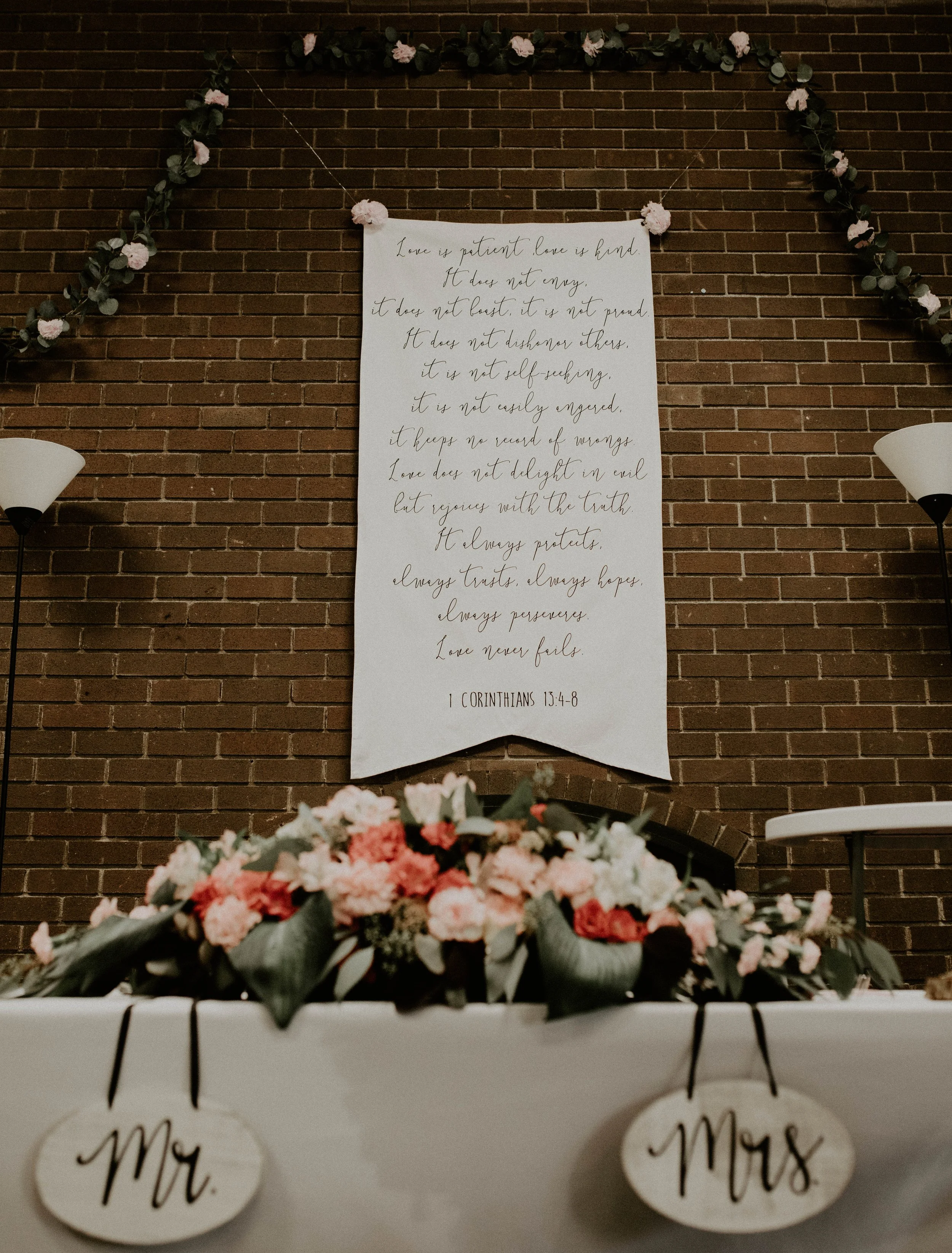 A large white banner with a Bible quote from 1 Corinthians 13:4-8 hangs on a brick wall, decorated with pink and white flowers and greenery. In the foreground, there is a table with floral arrangements and two wooden signs that say 'Mr.' and 'Mrs.' S
