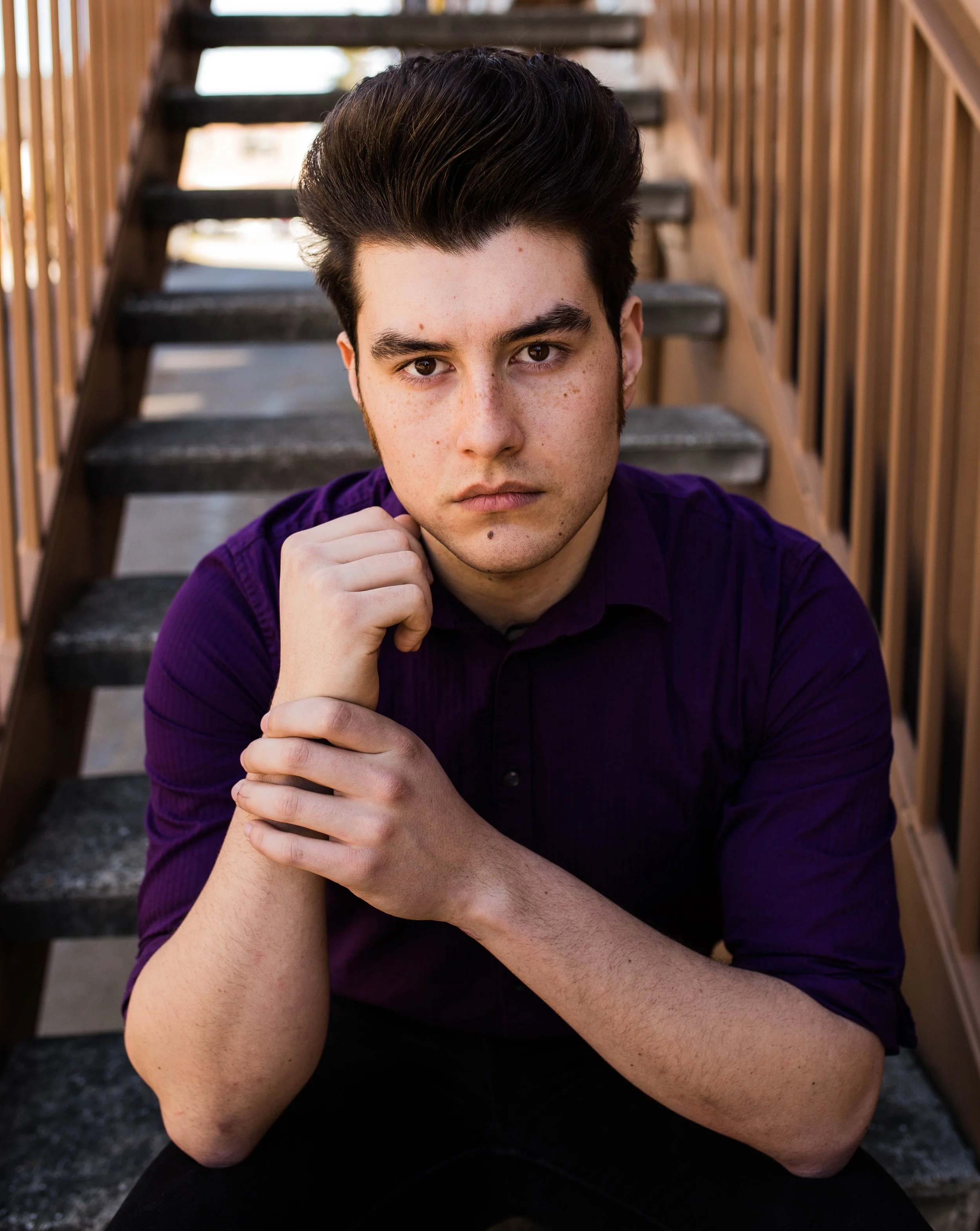 Young man with dark hair and freckles, wearing a purple shirt, sitting on outdoor stairs, looking directly at the camera with a serious expression. Seattle professional head shot photography