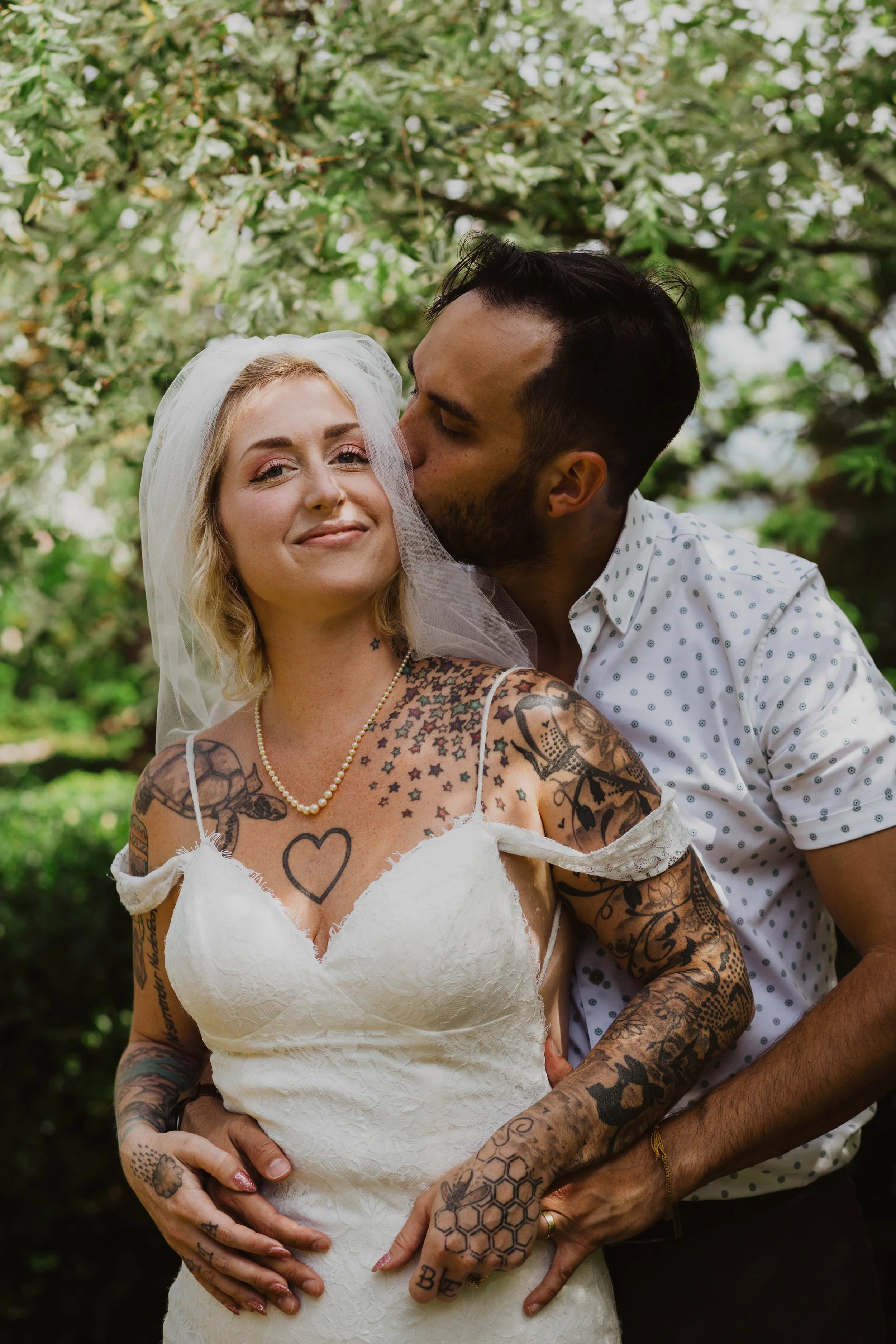 A woman with tattoos in a wedding dress has a man kissing her cheek from behind. They are outdoors with green foliage in the background. Seattle, WA wedding photography.