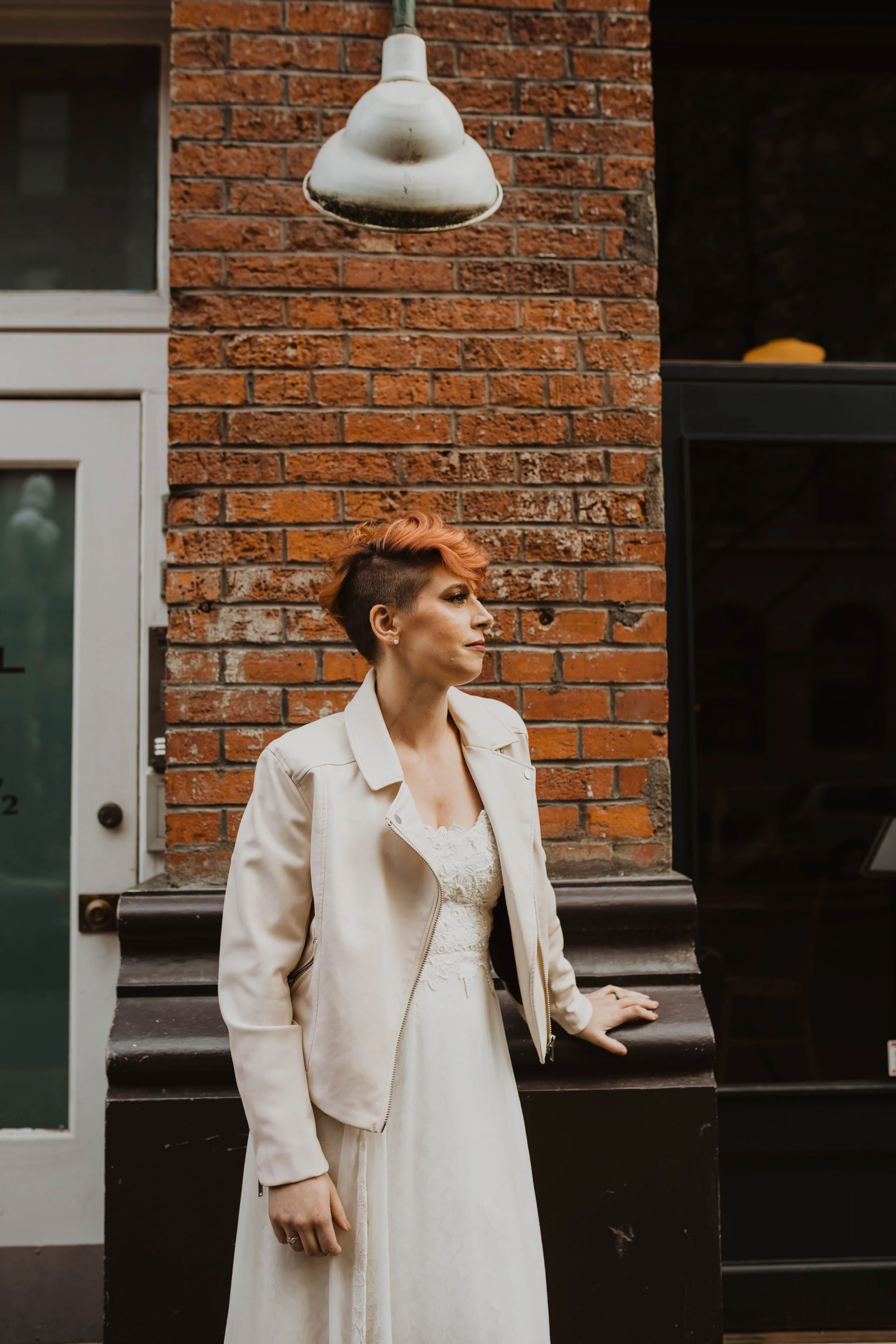 A woman with short, styled red hair and a white dress leaning against a black ledge in front of a brick wall, with a white door to her right and an outdoor lamp hanging above her. Pioneer Square, Seattle, WA wedding photography.