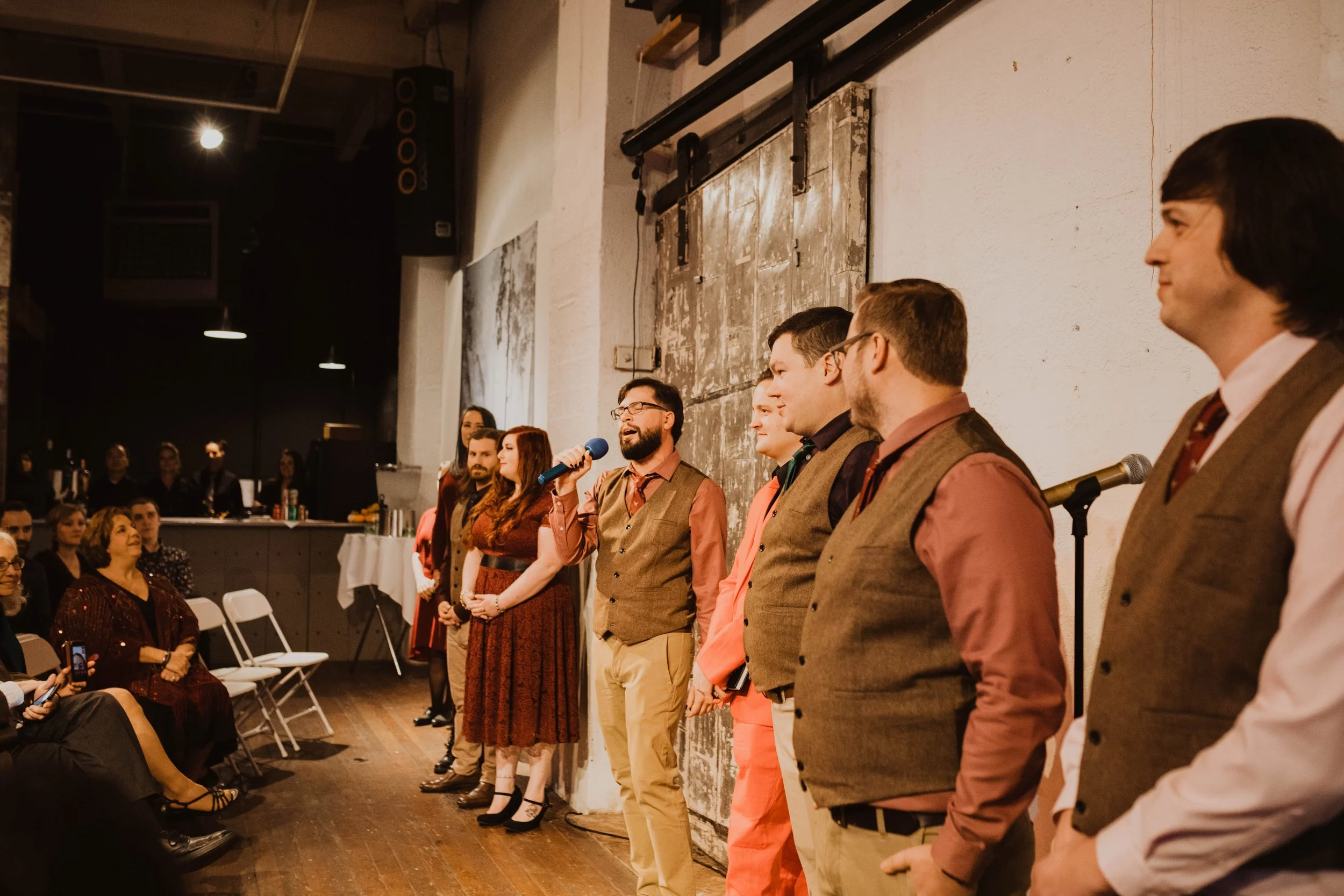 Group of people in vintage clothing standing along a textured wall at an indoor event, with some seated audience members in the foreground. Pioneer Square, Seattle, WA wedding photography.