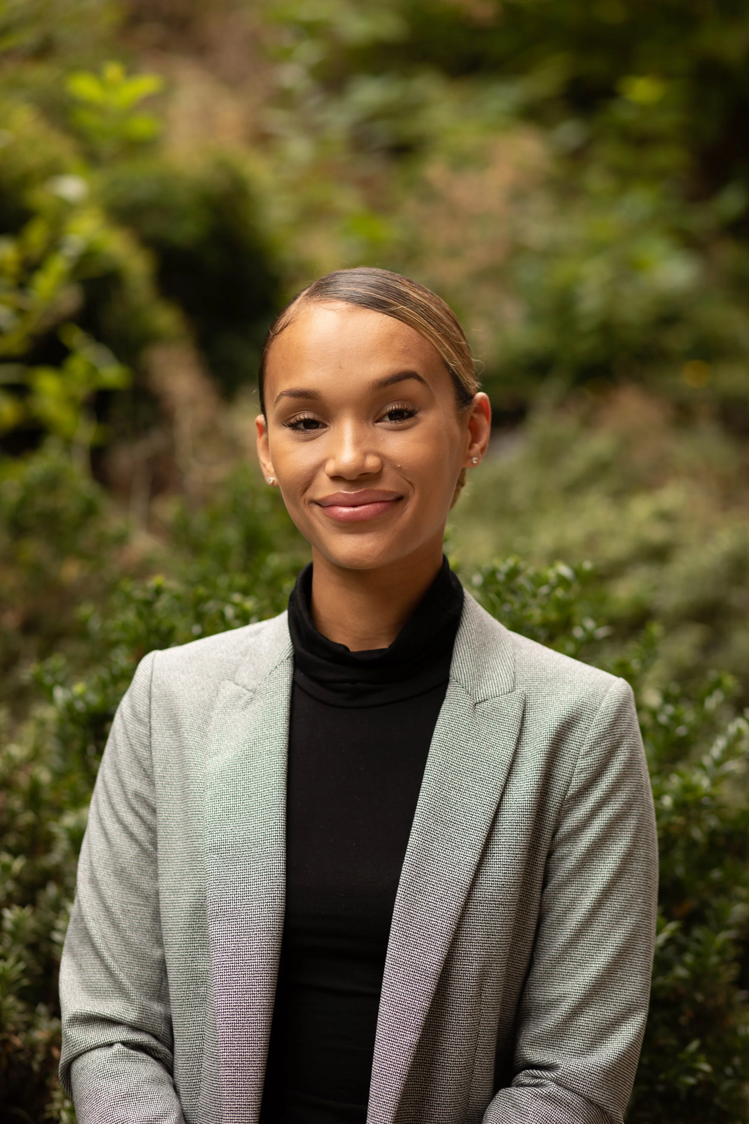 A young woman with light brown hair styled back, wearing a black turtleneck and a light gray blazer, smiling outdoors with blurred green foliage in the background. Seattle professional head shot photography