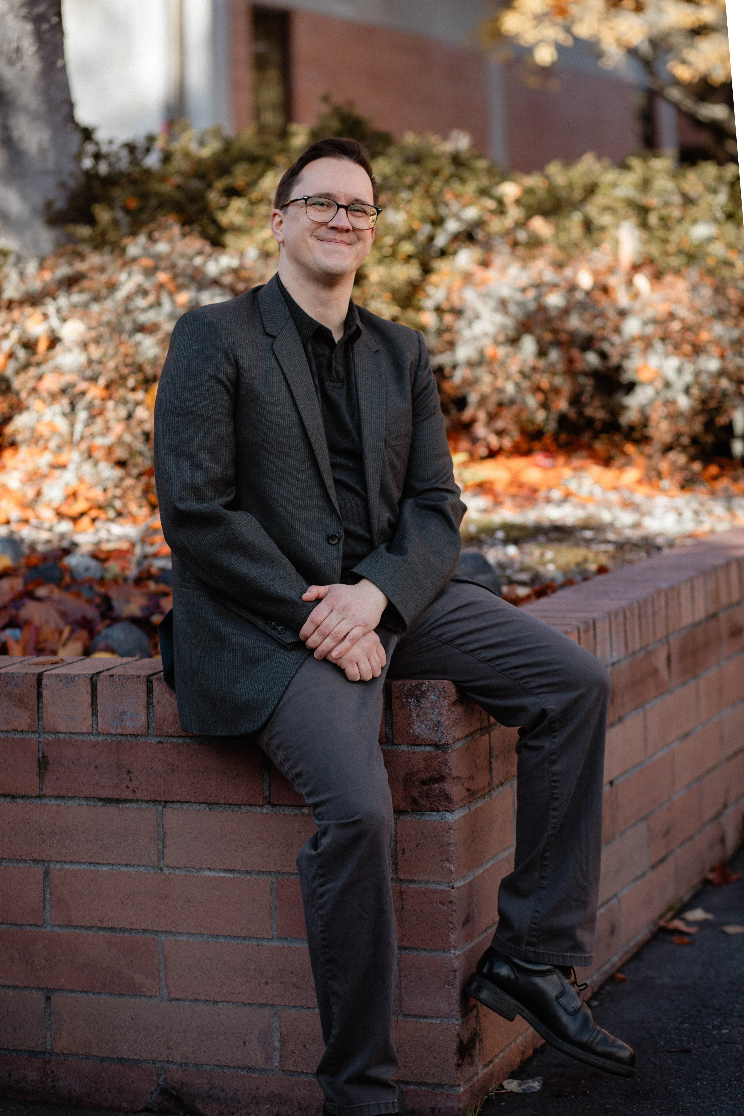 A man in a dark suit sitting on a brick wall outdoors with autumn foliage in the background. Seattle professional head shot photography
