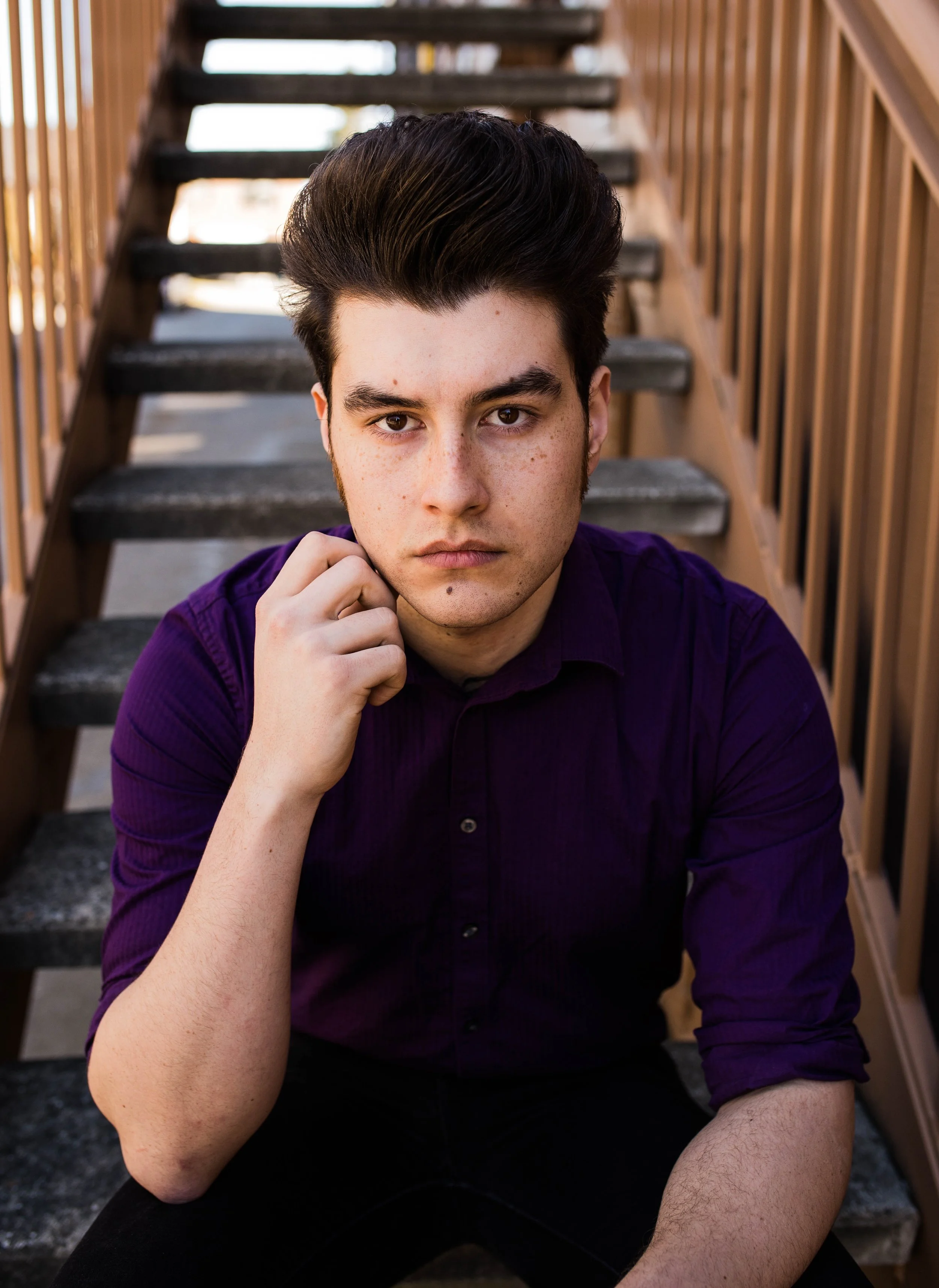 A young man with dark hair and freckles sitting on outdoor stairs, wearing a purple shirt and looking directly at the camera. Seattle professional head shot photography