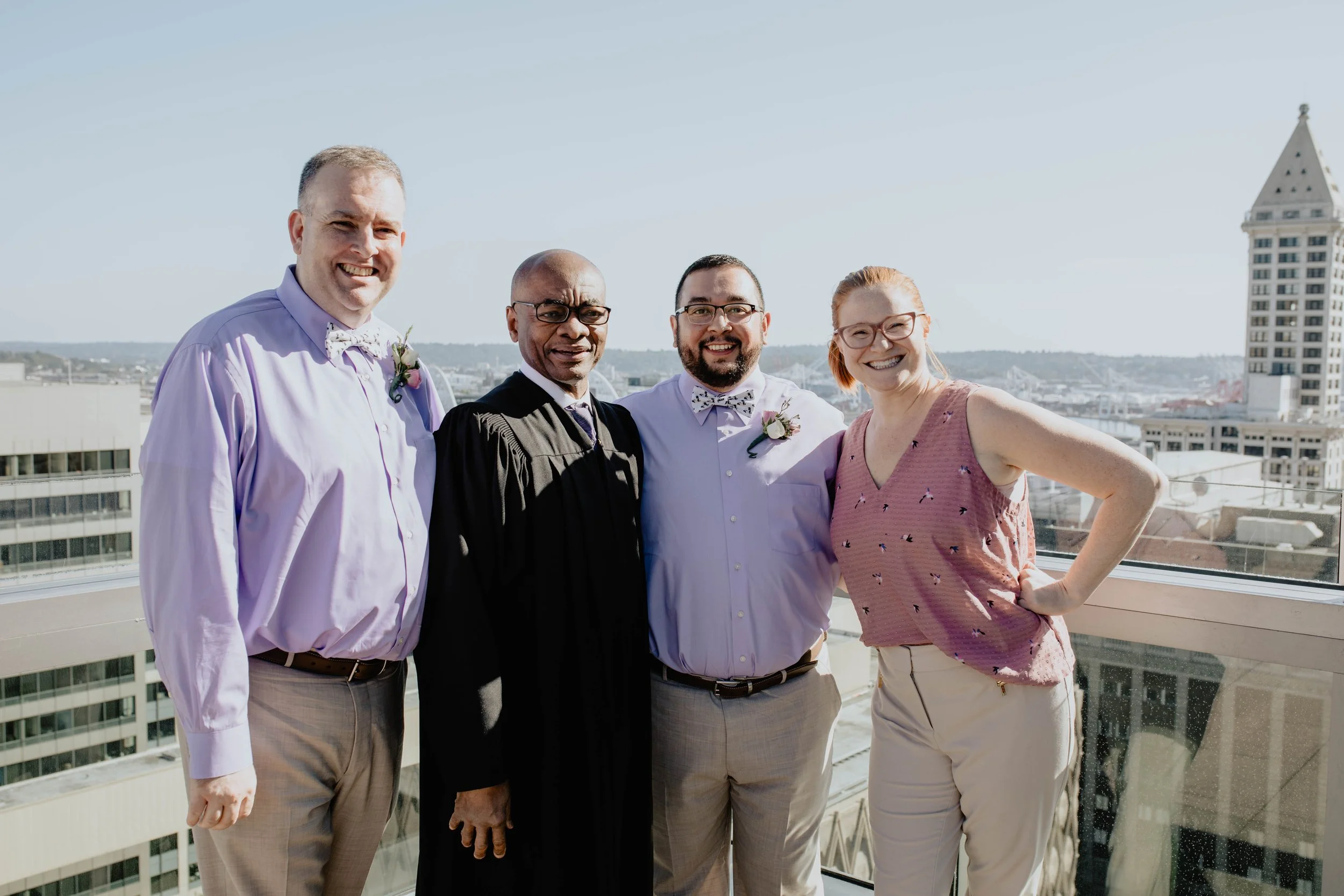 Four people celebrating a wedding on a rooftop with cityscape background, two men in dress shirts and bow ties, one person in a judge's robe, and a woman in a sleeveless top. Seattle Municipal Courthouse wedding photography.