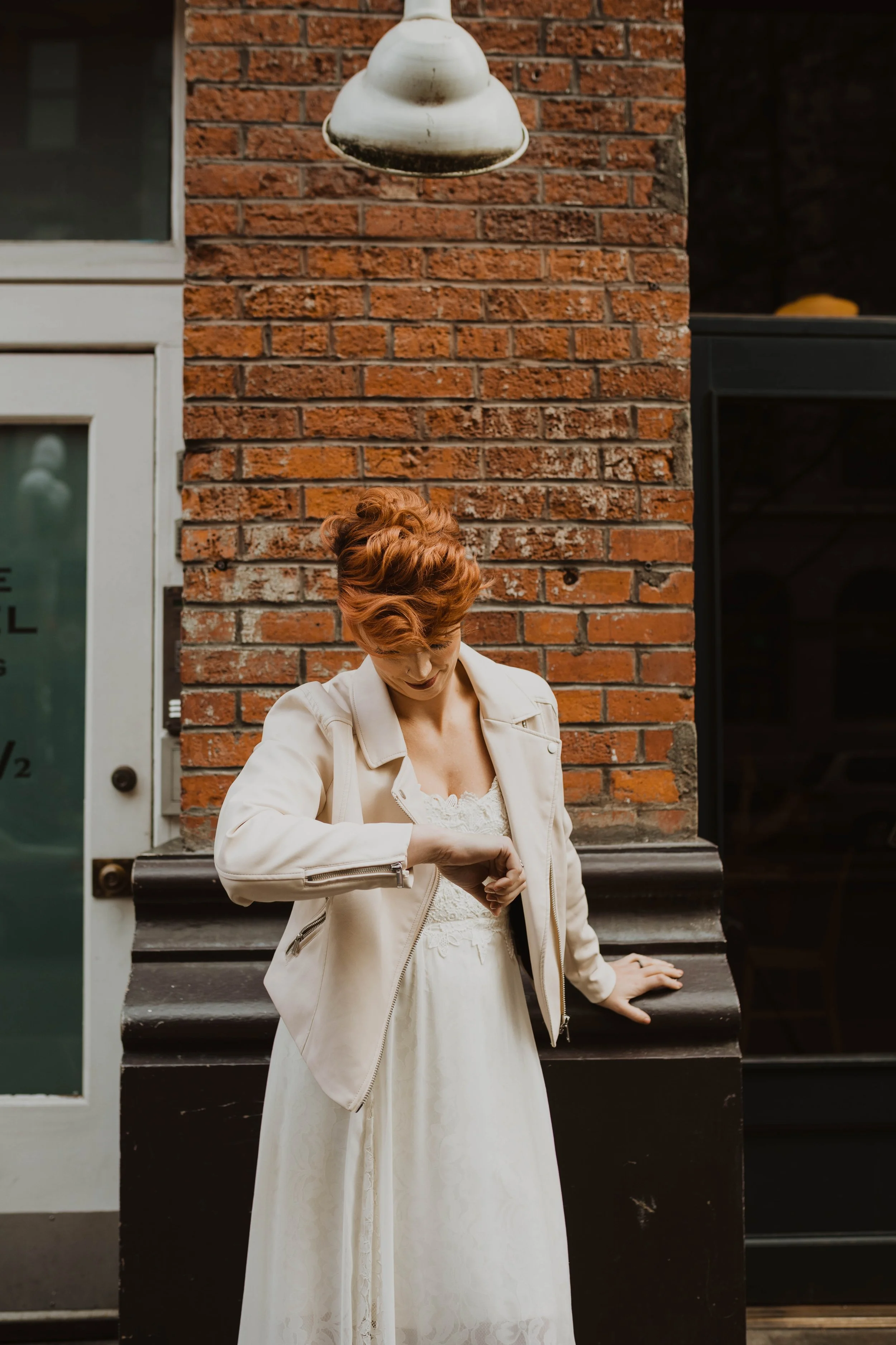A woman with red hair in a vintage hairstyle, wearing a white dress and a cream-colored leather jacket, looking at her watch while standing outside against a brick wall. Pioneer Square, Seattle, WA wedding photography.