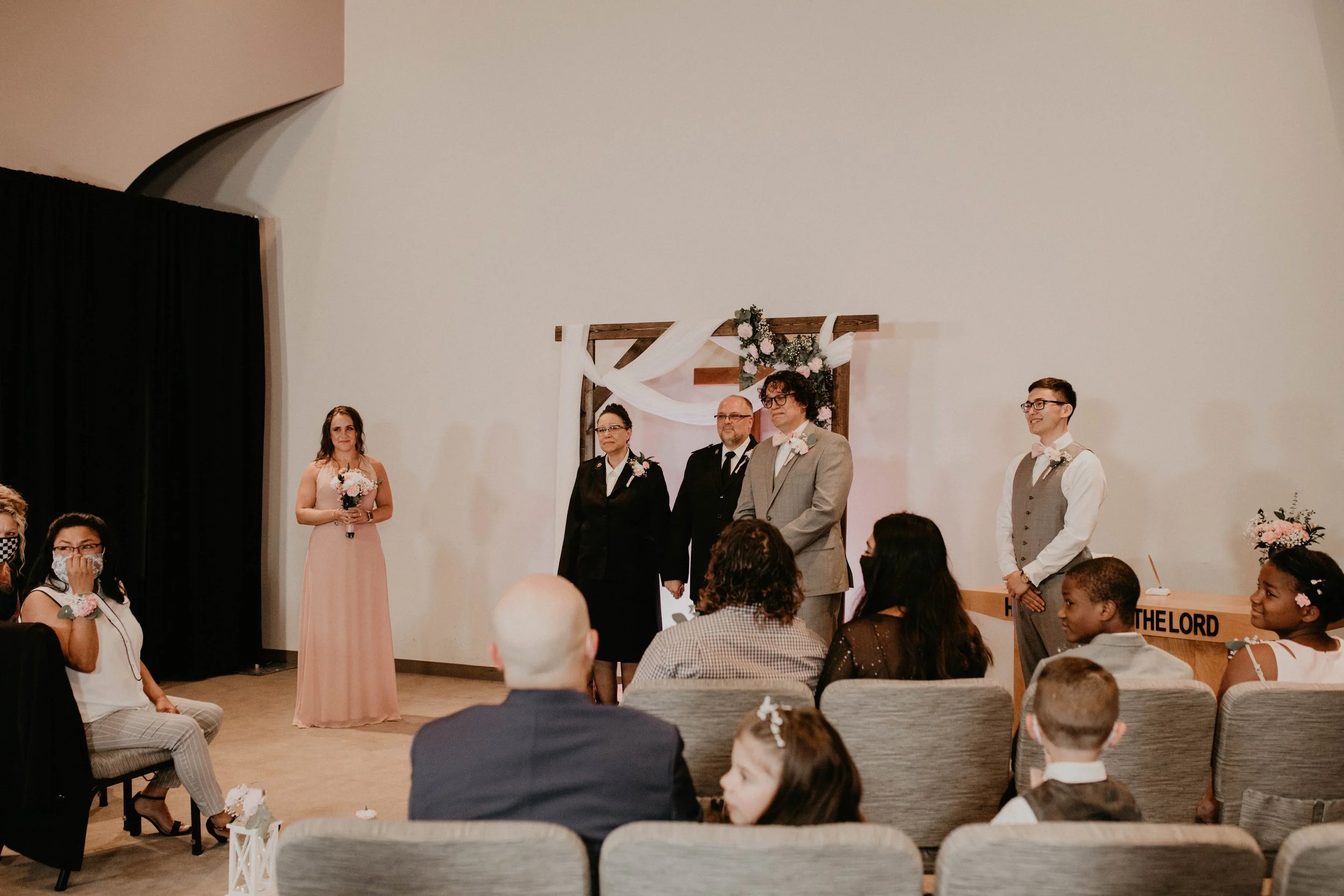 Wedding ceremony with three people standing at the altar, two women and one man, dressed in formal attire, with guests seated in front. The background includes a decorative wooden arch with white fabric and flowers. Seattle, WA wedding photography.