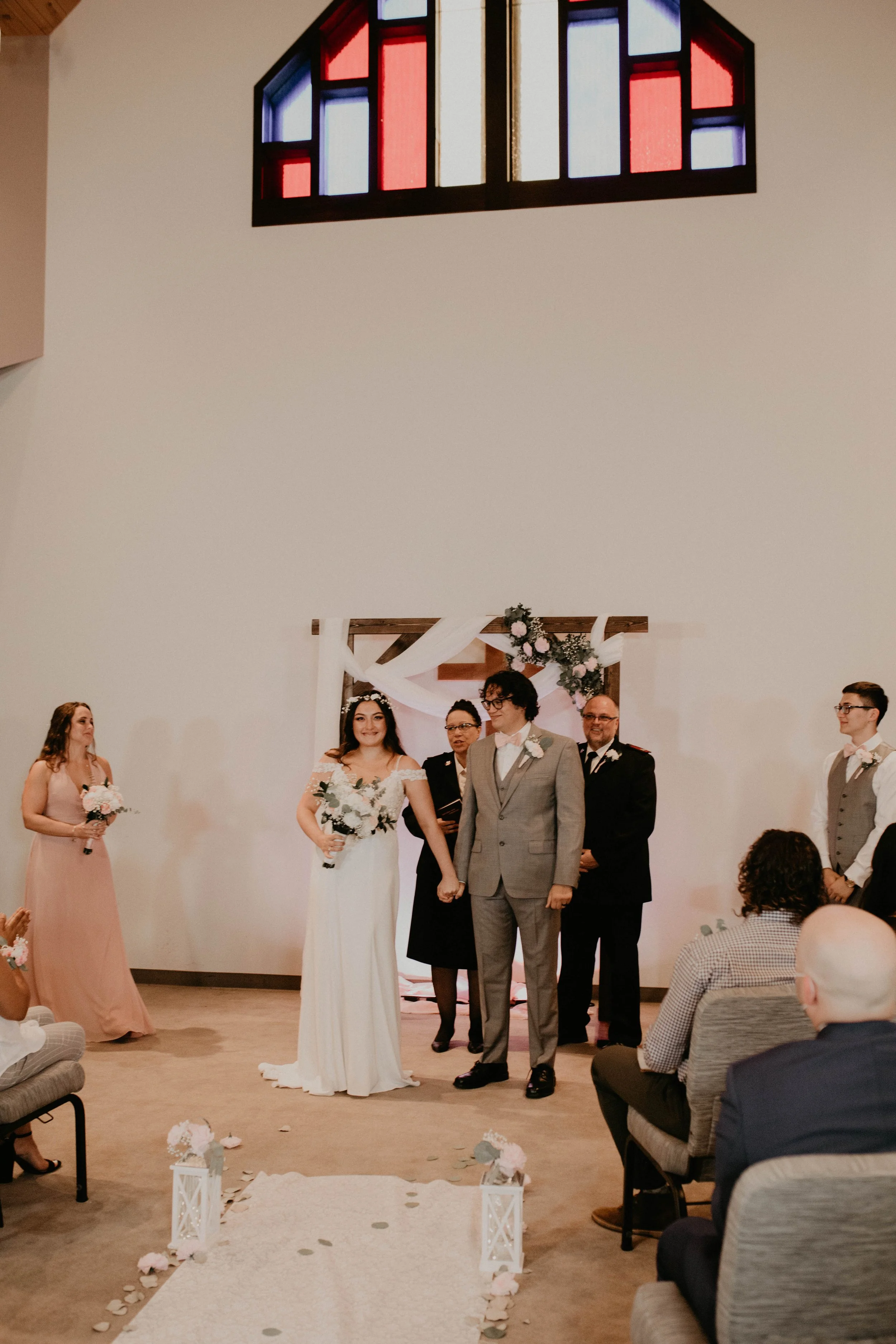 A wedding ceremony taking place inside a church with a bride, groom, officiant, and guests seated on chairs. The bride and groom are holding hands, and the bride is wearing a white dress while the groom is dressed in a gray suit. There's a floral arc
