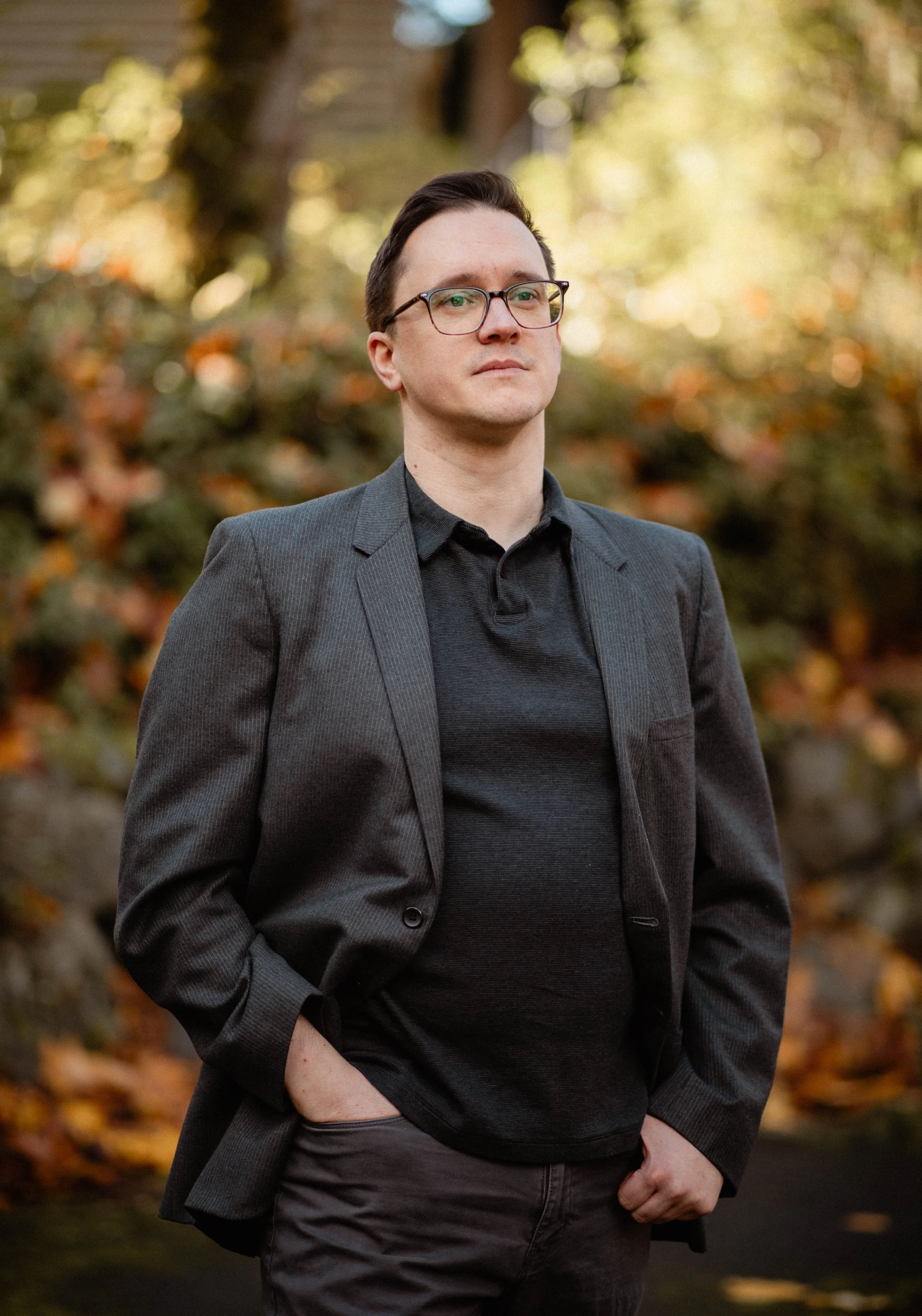 A man wearing glasses, a dark blazer, and a black shirt standing outdoors during fall with trees and fallen leaves in the background. Seattle professional head shot photography