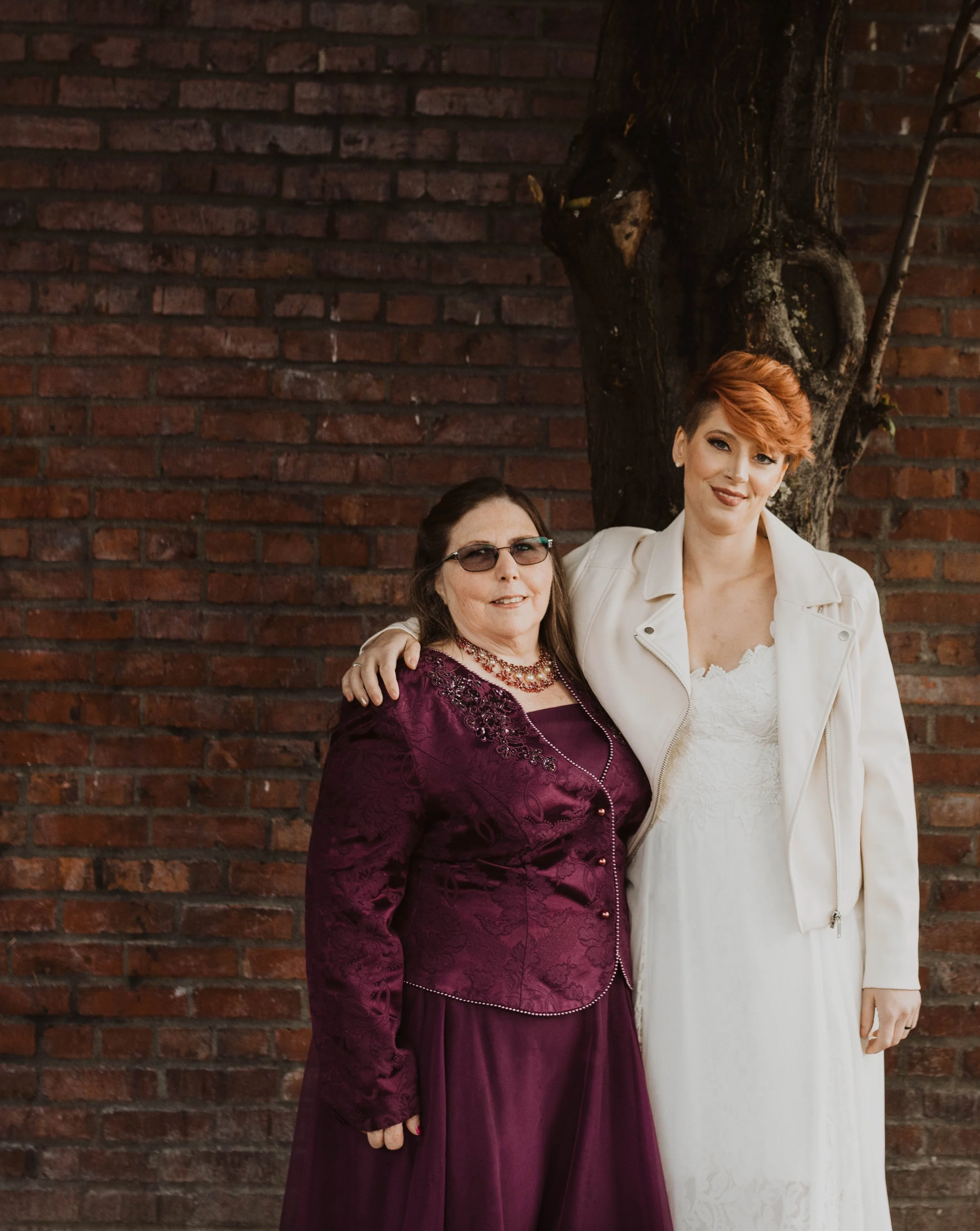 Two women standing close together with their arms around each other, posing outdoors in front of a brick wall and a tree. Pioneer Square, Seattle, WA wedding photography.