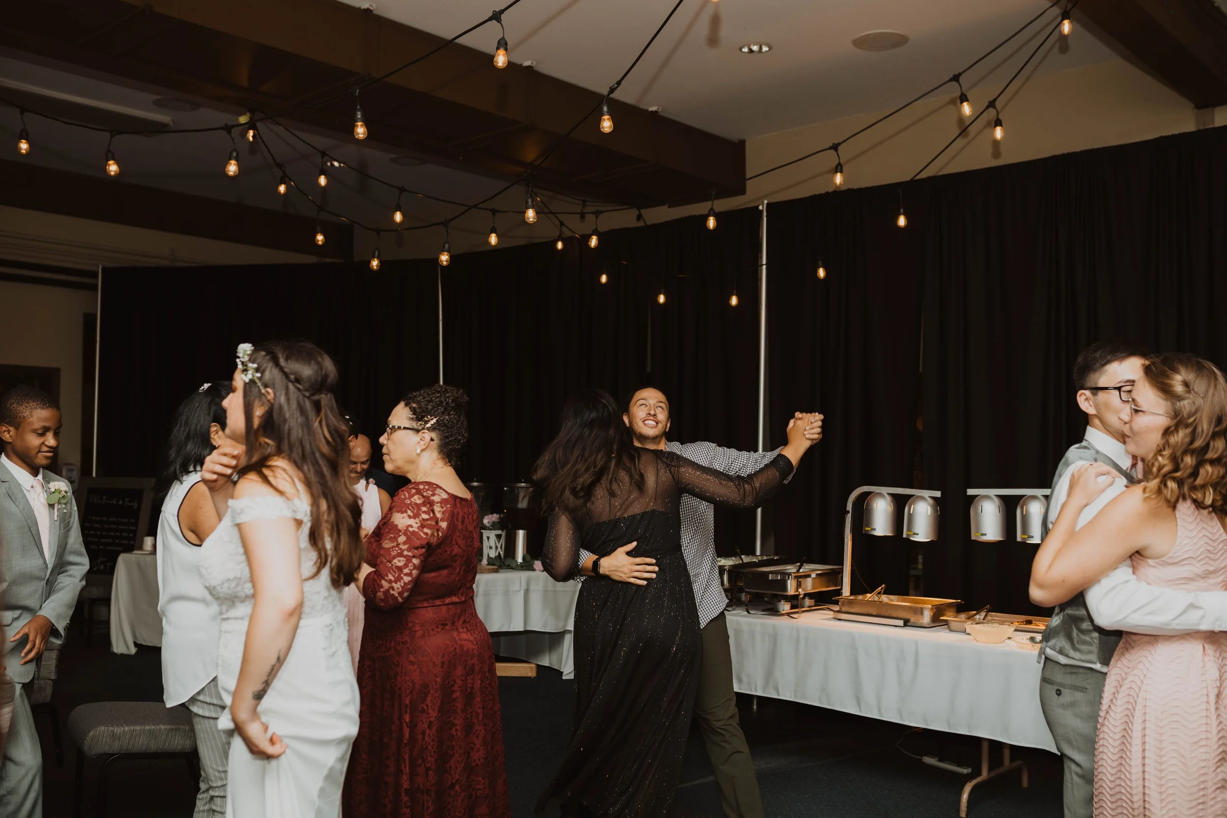 People dancing and socializing at a wedding reception, with string lights hanging from the ceiling and buffet table in the background. Seattle, WA wedding photography.