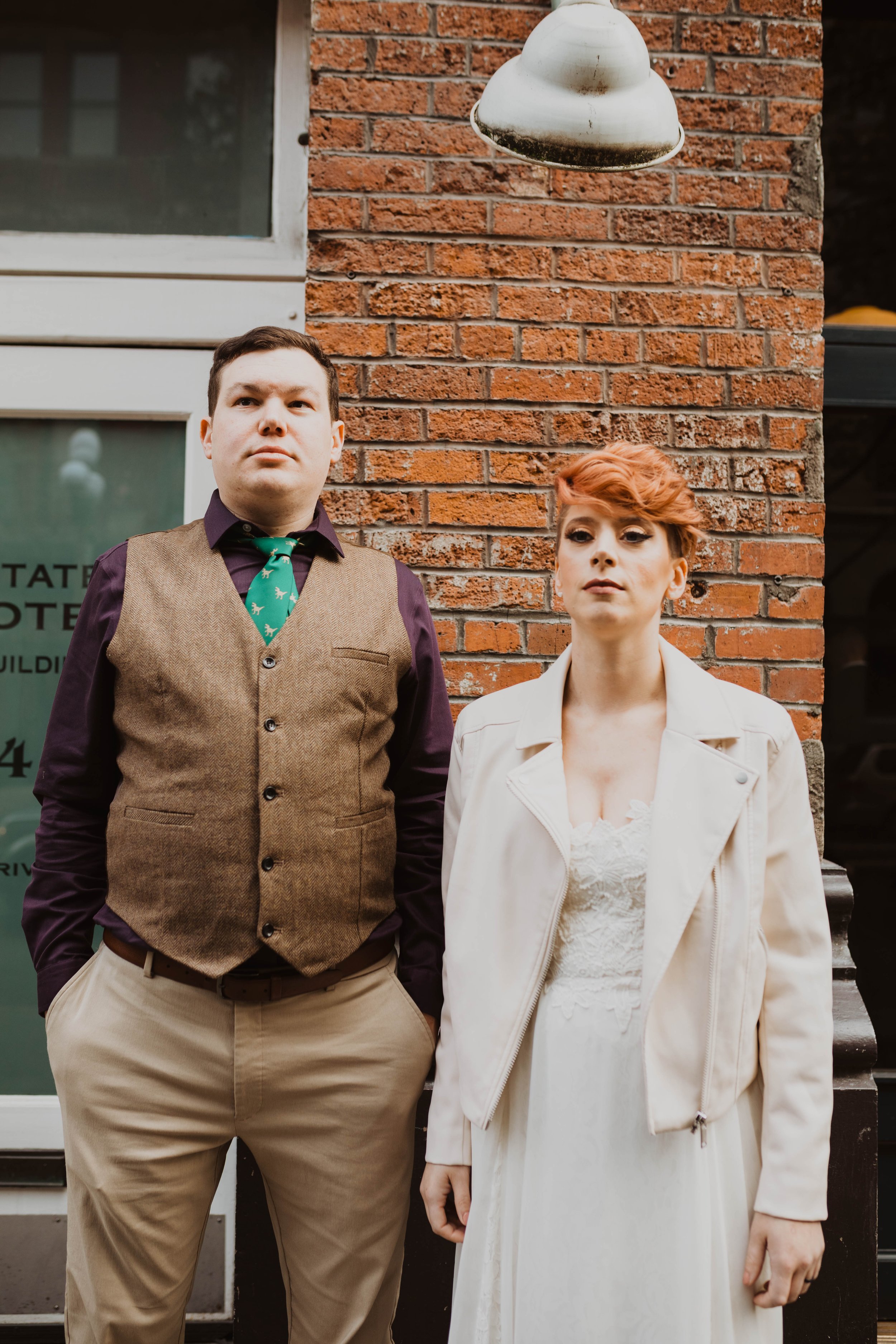 A man and woman standing against a red brick wall, with a white window and an old-fashioned outdoor light fixture above them. Pioneer Square, Seattle, WA wedding photography.