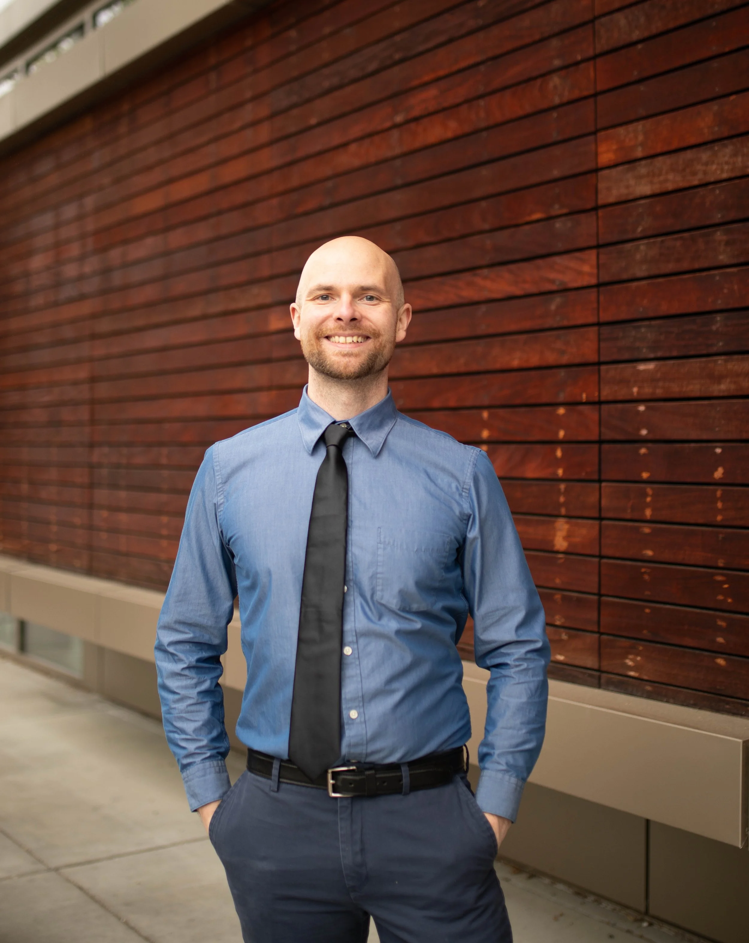 A man with a beard, wearing a blue dress shirt, black tie, and dark pants, standing outside in front of a wooden wall, smiling with his hands in his pockets. Seattle professional head shot photography