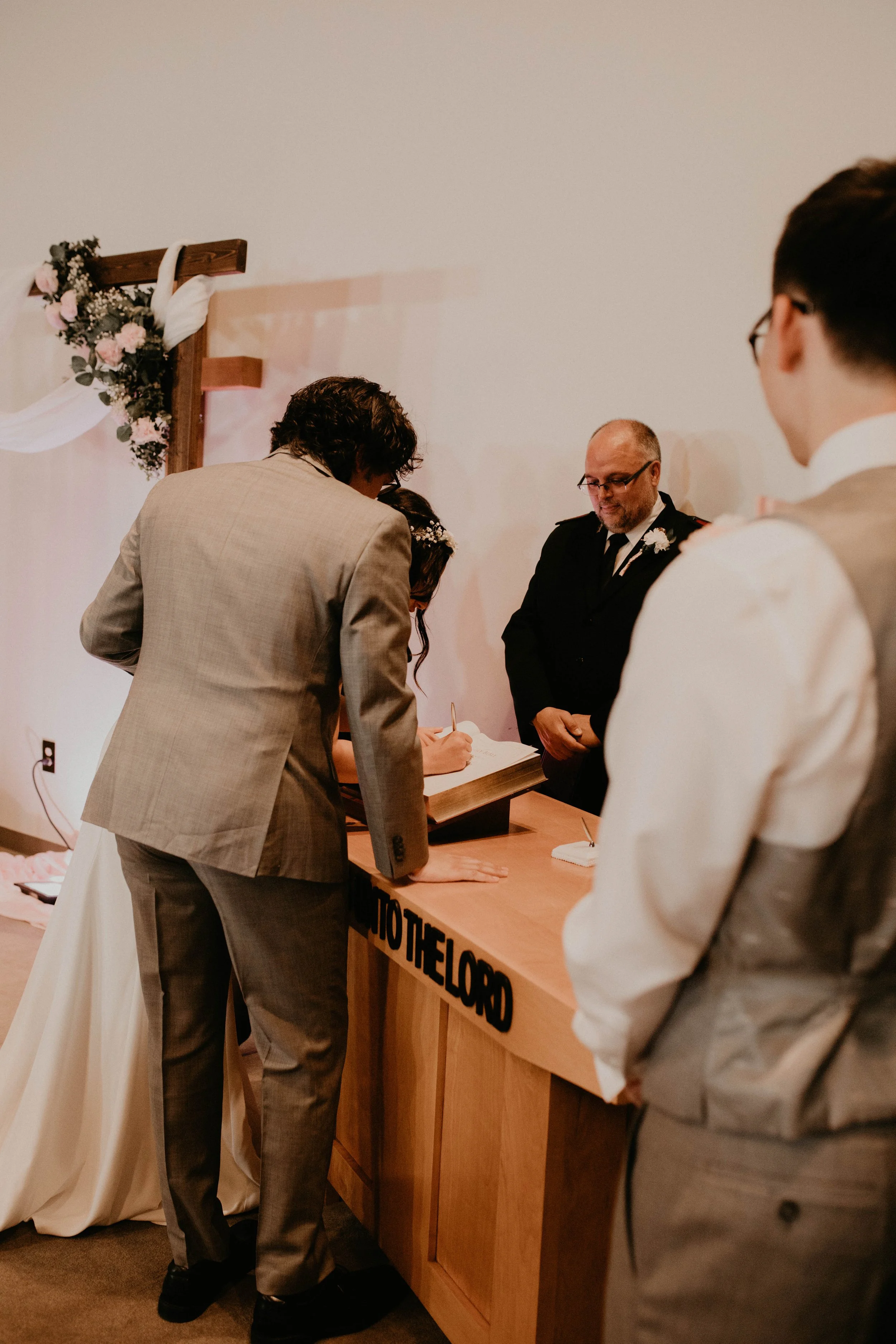 A wedding ceremony with a couple signing documents at a wooden table with a floral background, attended by officiants and guests. Seattle, WA wedding photography.