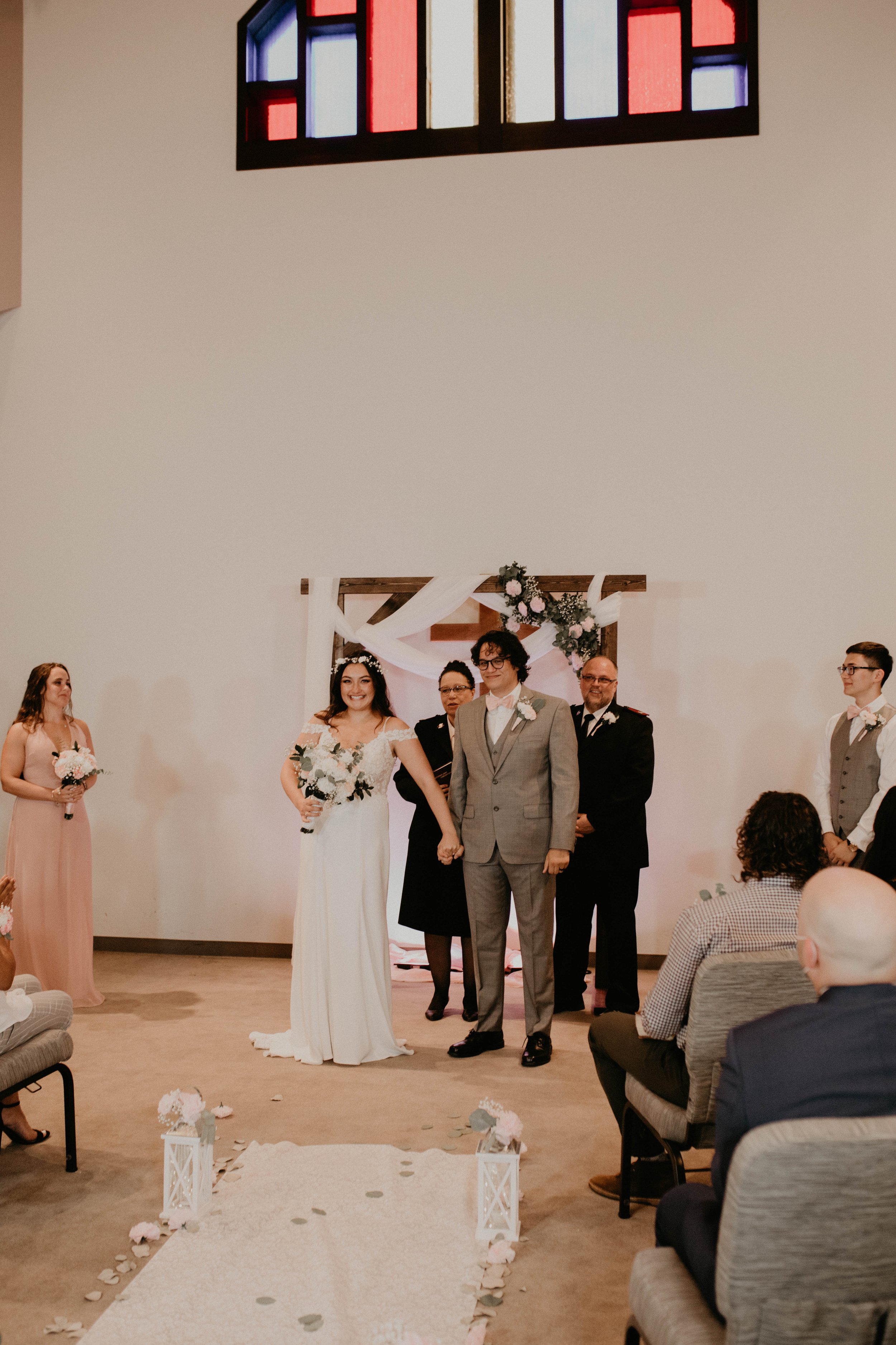 A wedding ceremony with a bride and groom holding hands in front of guests inside a church, with bridesmaids and groomsmen standing beside them, and a decorated altar behind. Seattle, WA wedding photography.