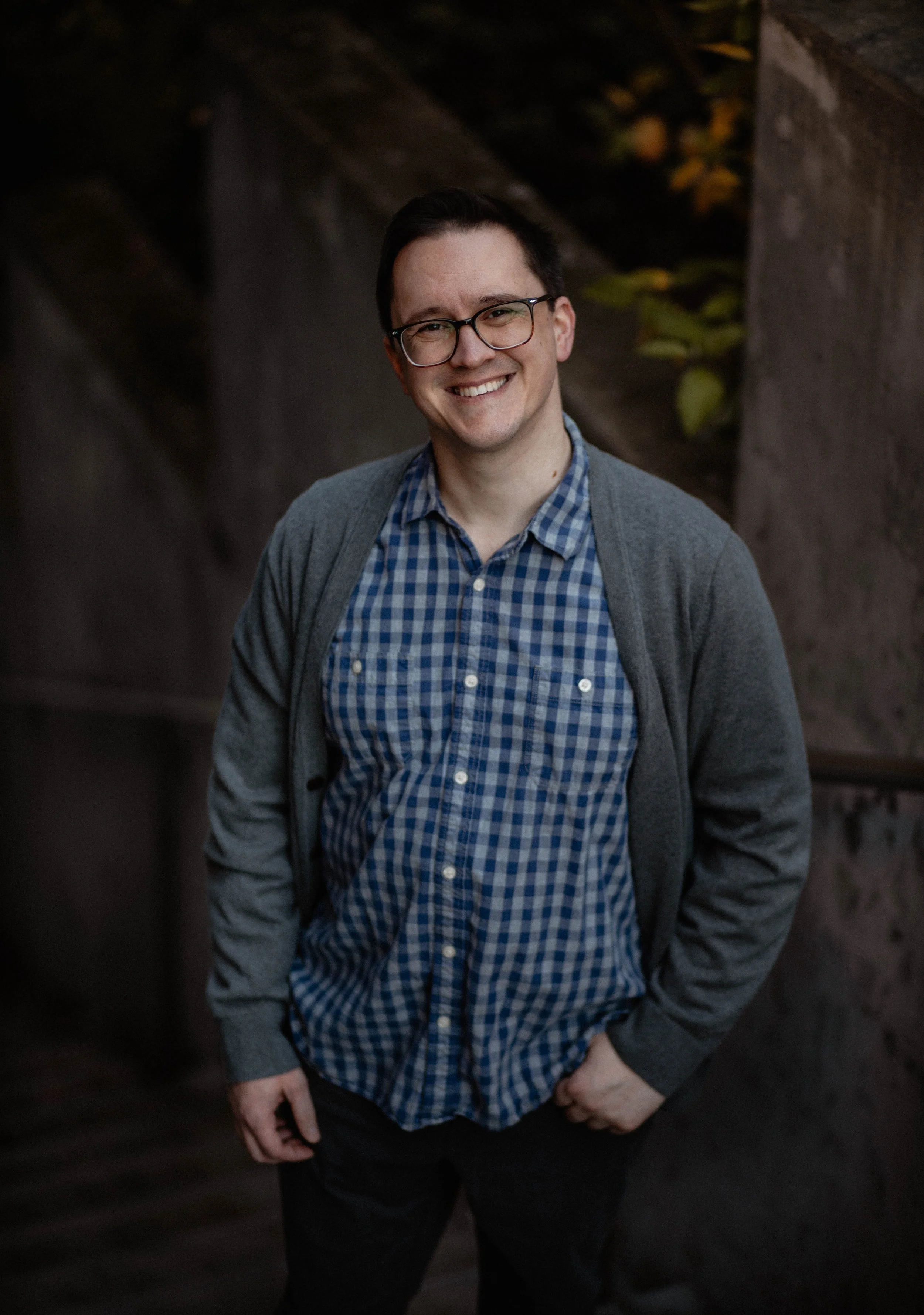 A young man with glasses, wearing a gray cardigan over a blue checkered shirt, smiling outdoors near a stone wall with some autumn leaves in the background. Seattle professional head shot photography