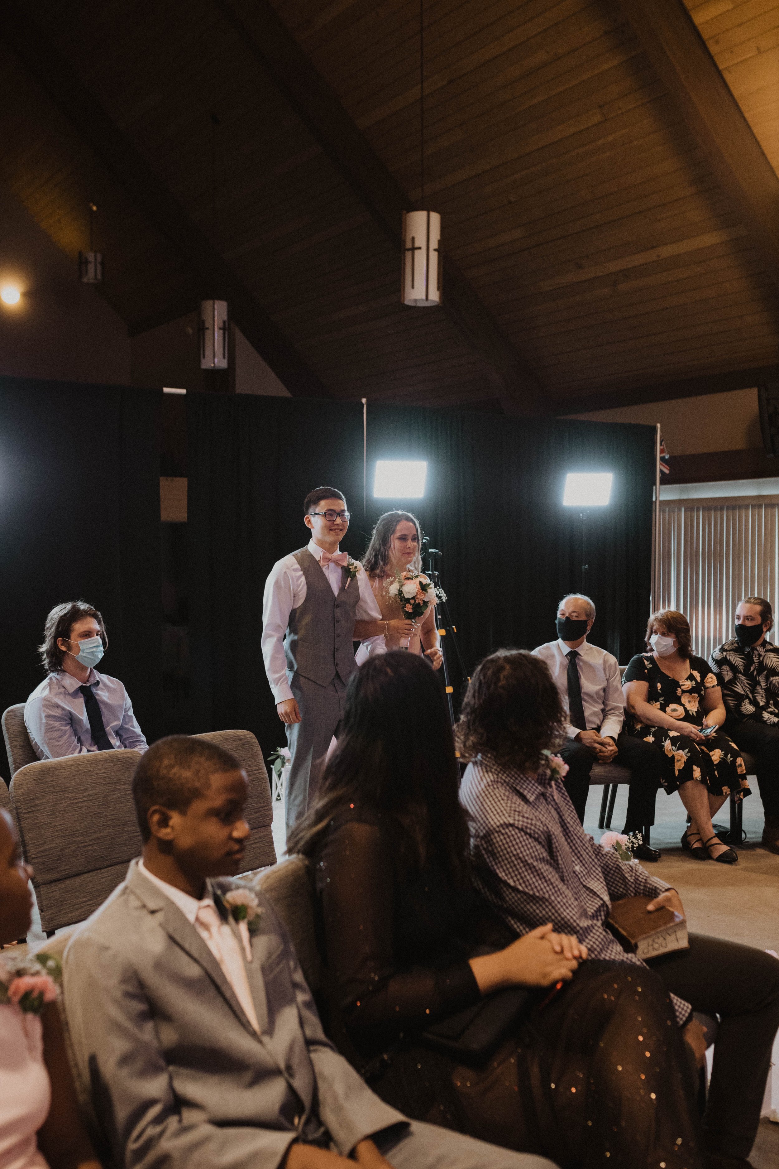 A wedding ceremony with a couple standing together, surrounded by seated guests. The couple is dressed in wedding attire; the woman holds a bouquet and the man is in a vest and tie. The guests are wearing masks, seated attentively, some with books or
