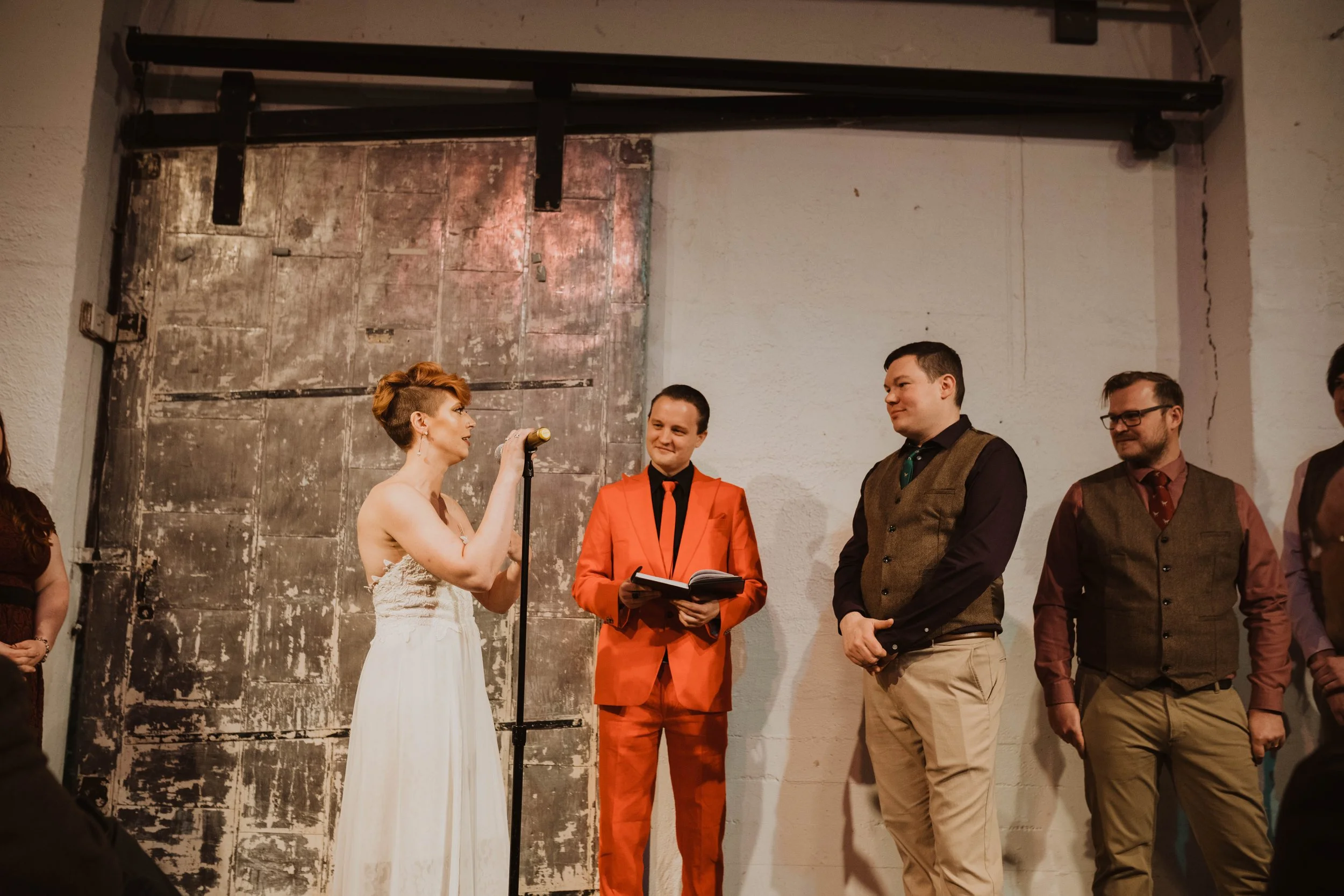 A woman in a white wedding dress speaking into a microphone during a wedding ceremony, with three men standing nearby, one in a red suit, and the others in brown vests and dress shirts, in front of a rustic backdrop.