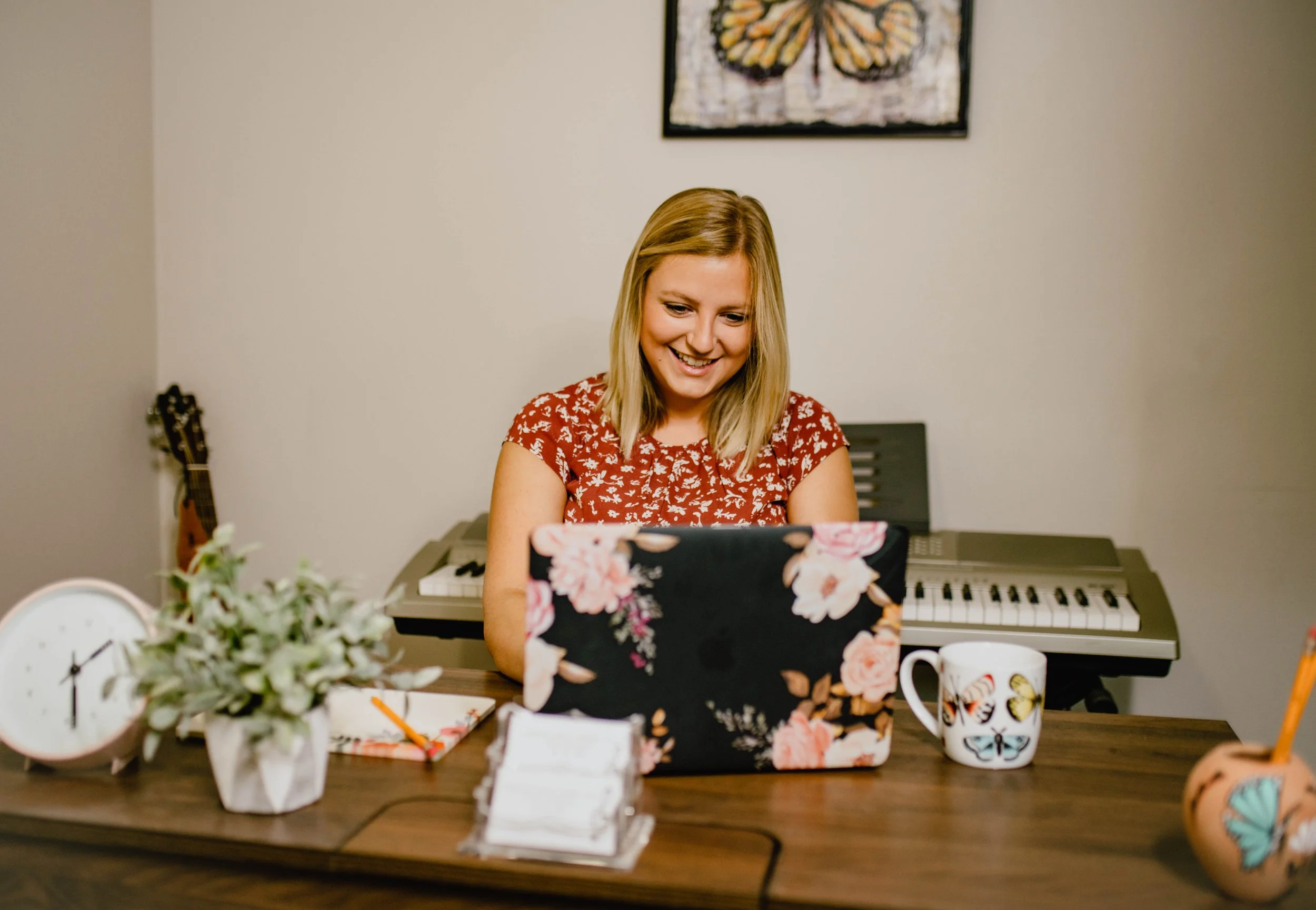 A smiling woman with blonde hair working on a laptop at a wooden desk, with a keyboard, a clock, a potted plant, a mug with butterflies, and a notebook with a pen. In the background, a guitar and a painting of a butterfly on the wall.