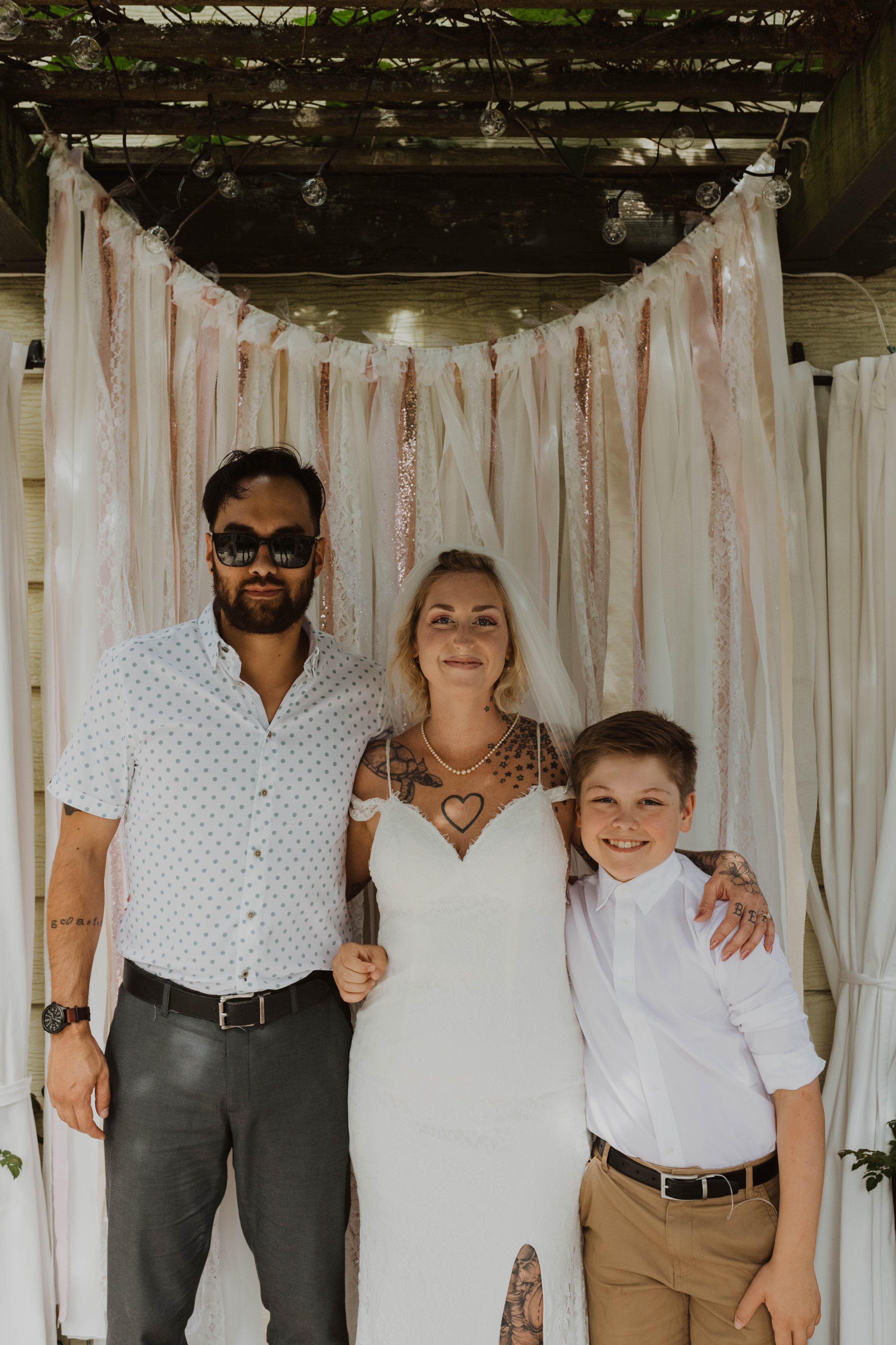 A bride with tattoos in a white wedding gown, standing between a man with sunglasses and a young boy, in front of a decorative backdrop with pink and white ribbons and string lights. Seattle, WA wedding photography.