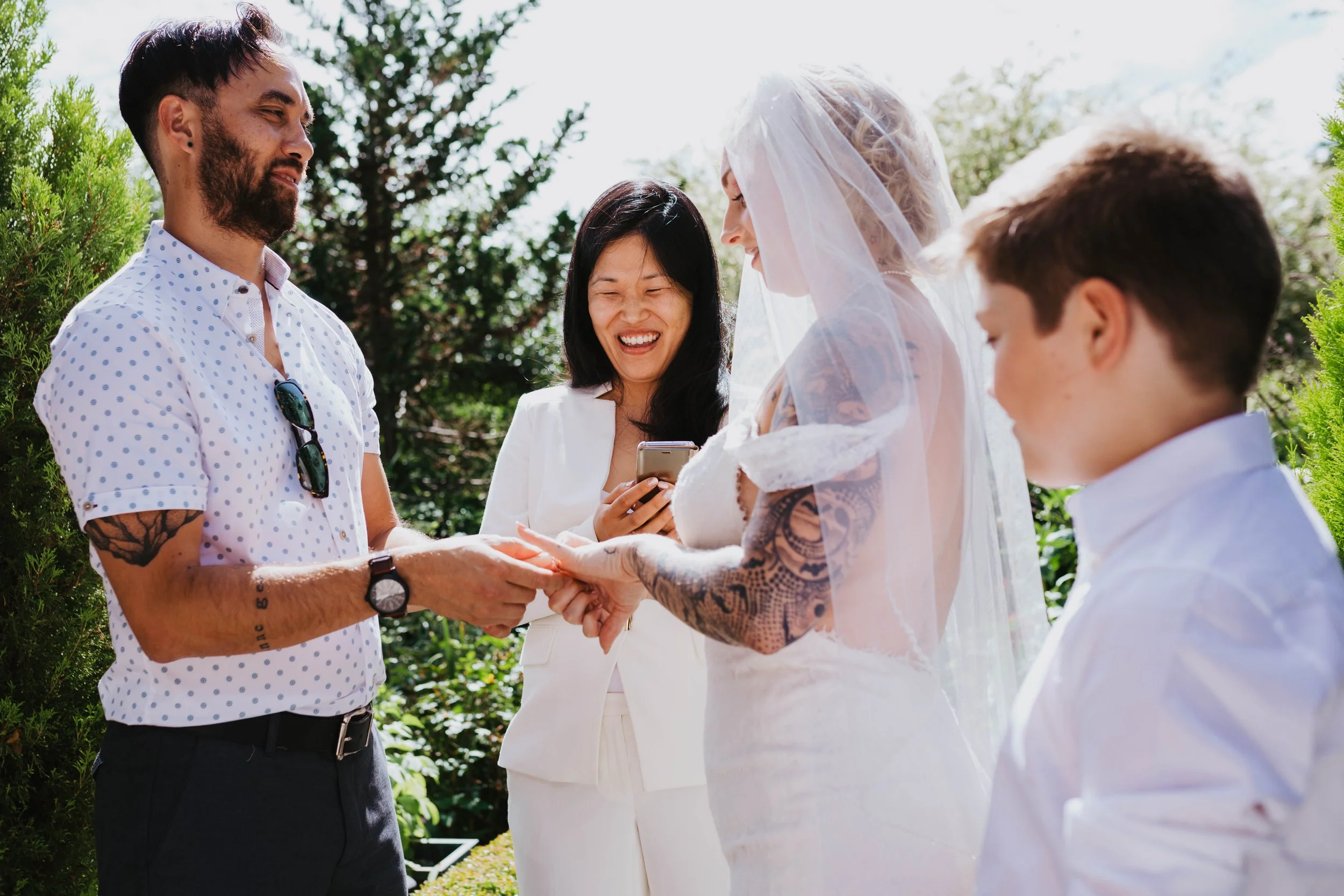 A couple getting married outdoors, with the bride in a white dress and veil, exchanging rings while an officiant, a woman in white, smiles and looks at them. A man and a boy, dressed in white shirts, stand nearby, and lush greenery surrounds them.