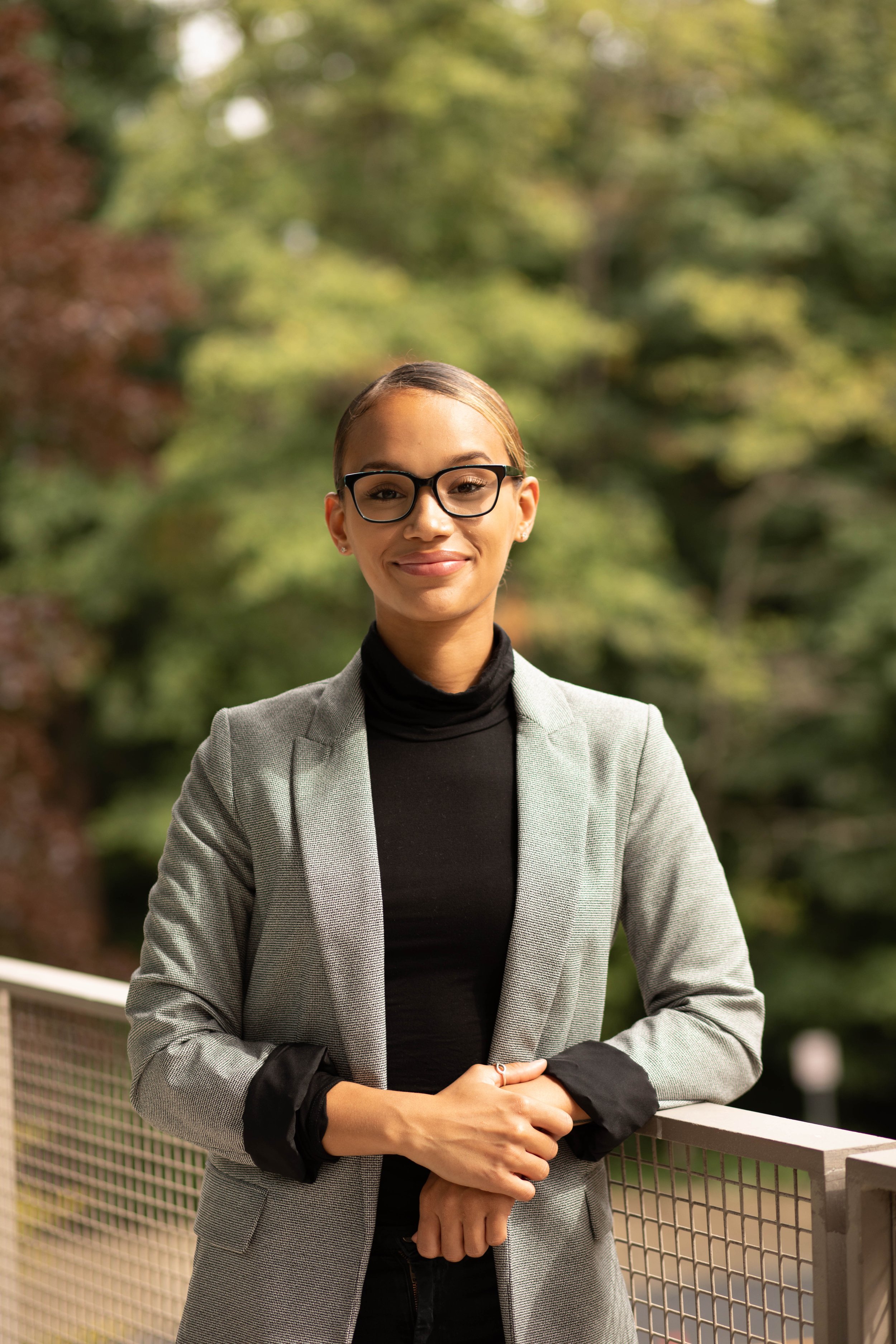 A young woman with glasses, wearing a gray blazer and black turtleneck, stands outdoors with green trees in the background, smiling softly at the camera. Seattle professional head shot photography