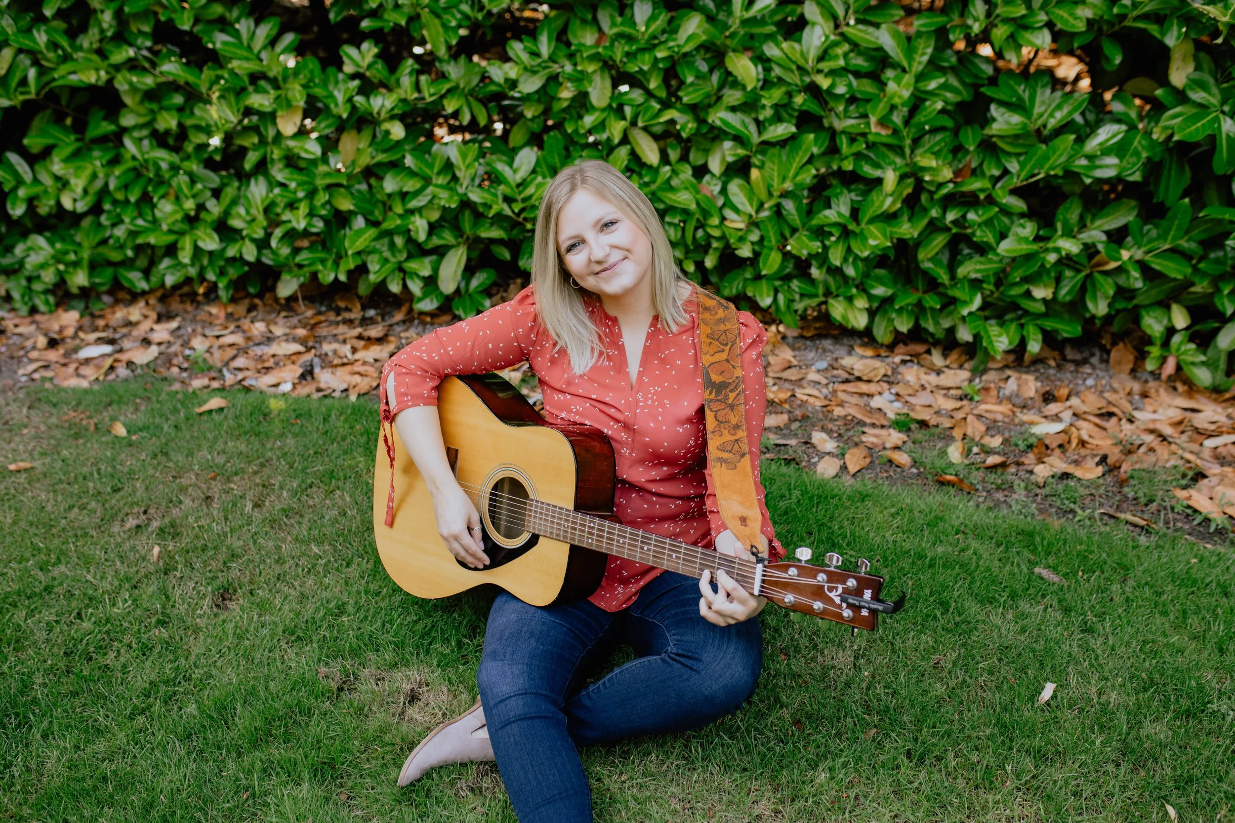 A young woman with blonde hair sitting on green grass, holding an acoustic guitar, smiling, in front of green bush with brown leaves on the ground. Seattle professional head shot photography