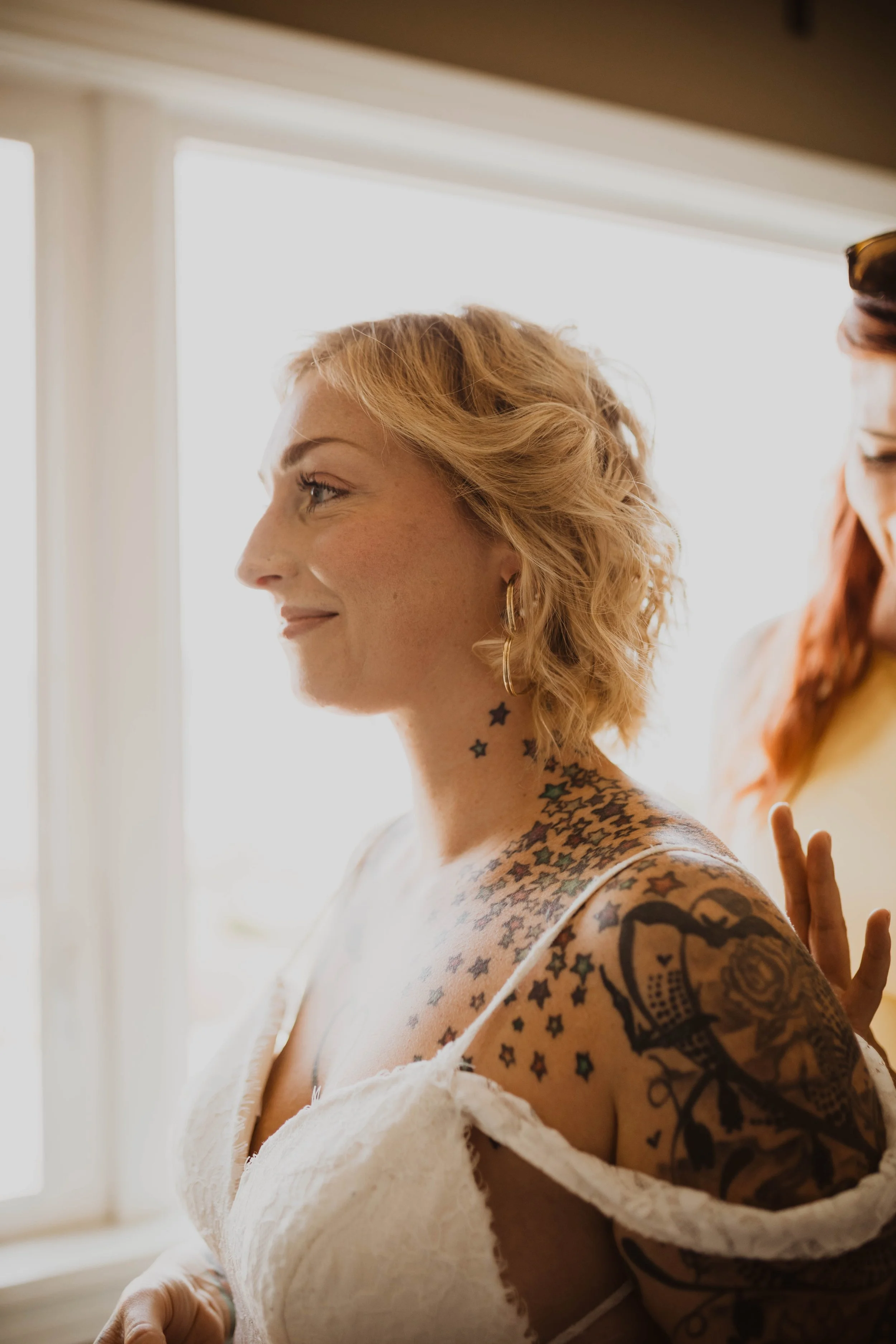 A woman with short, curly blonde hair and tattoos on her neck and shoulder, smiling slightly. She is wearing gold hoop earrings and a white, strapless top. Natural light illuminates her face from behind, creating a bright background. Seattle, WA wedd