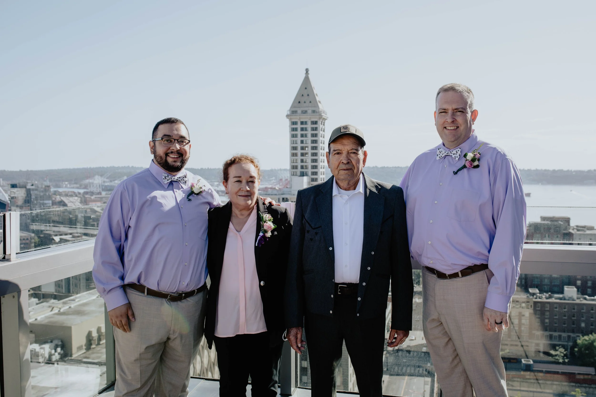 Family portrait on a rooftop with city skyline in the background, four people smiling, two men in matching shirts and bow ties with boutonnieres, a woman and an older man standing between them. Seattle Municipal Courthouse wedding photography.