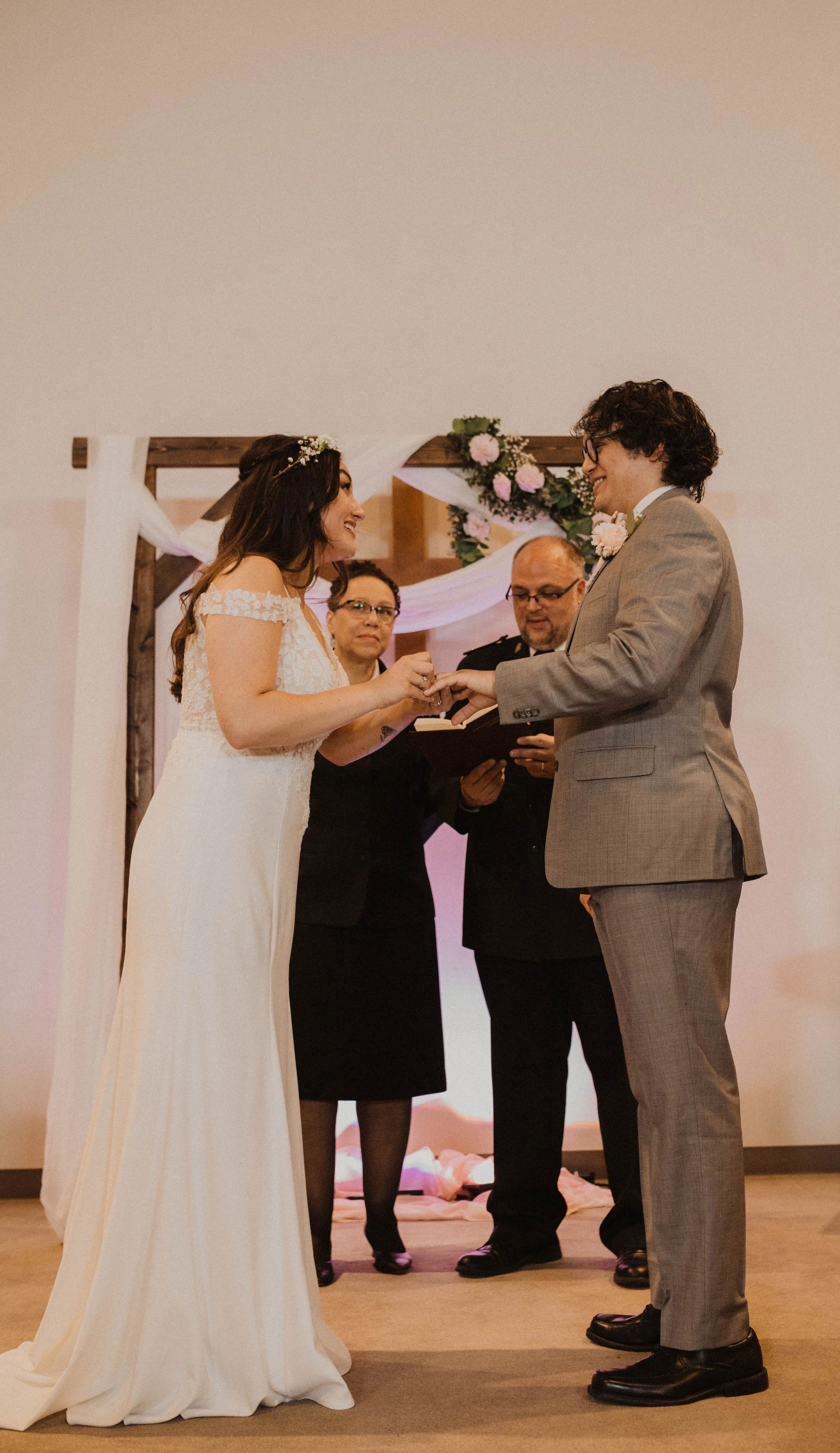 A bride and groom exchanging rings during their wedding ceremony, with two officiants standing behind them and decorated with pink and white flowers and white drapery. Seattle, WA wedding photography.