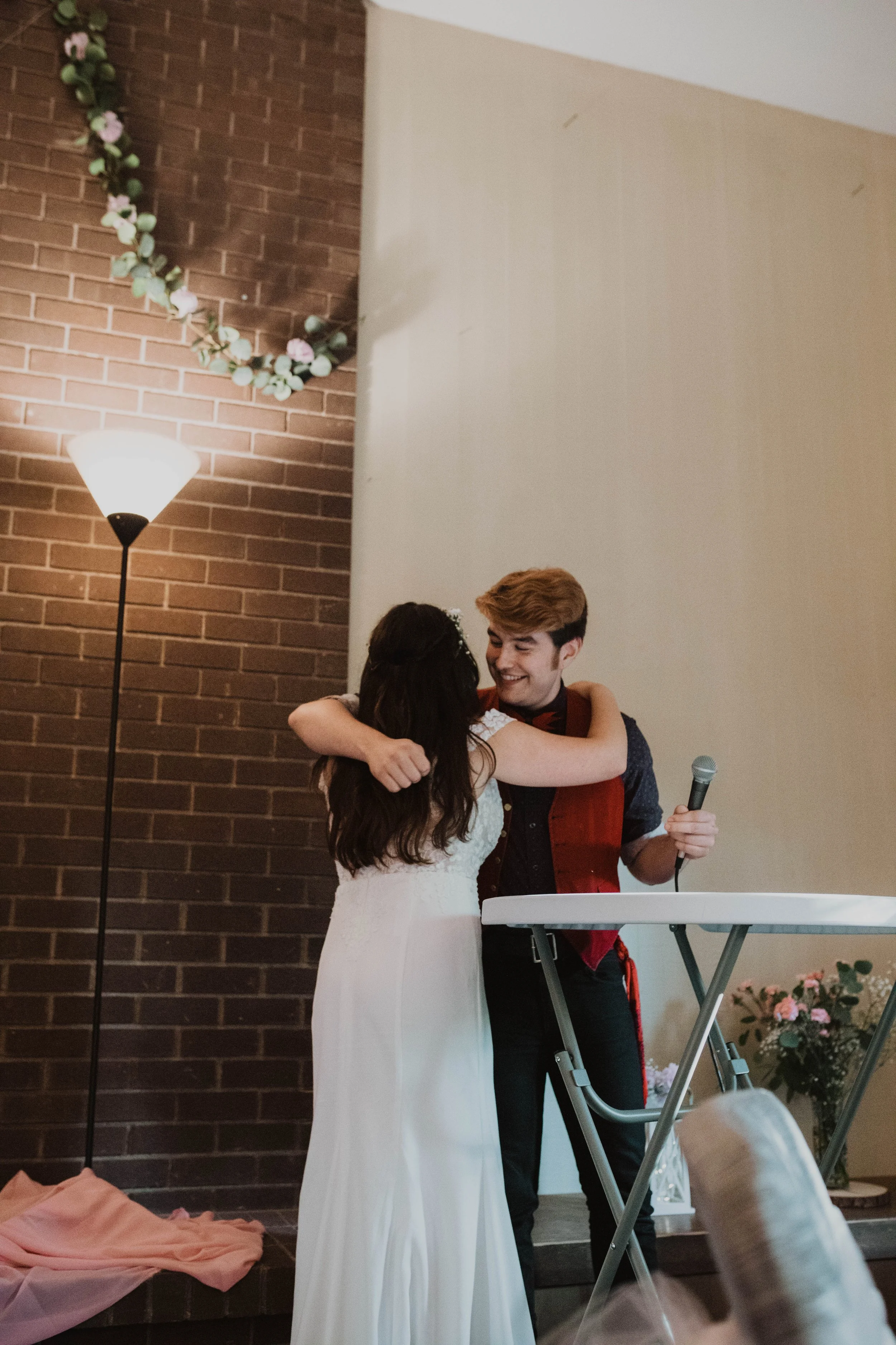 A couple hugging during a wedding ceremony, with the woman in a white dress and the man in dark clothes, holding a microphone, standing next to a table with flowers in the background. Seattle, WA wedding photography.
