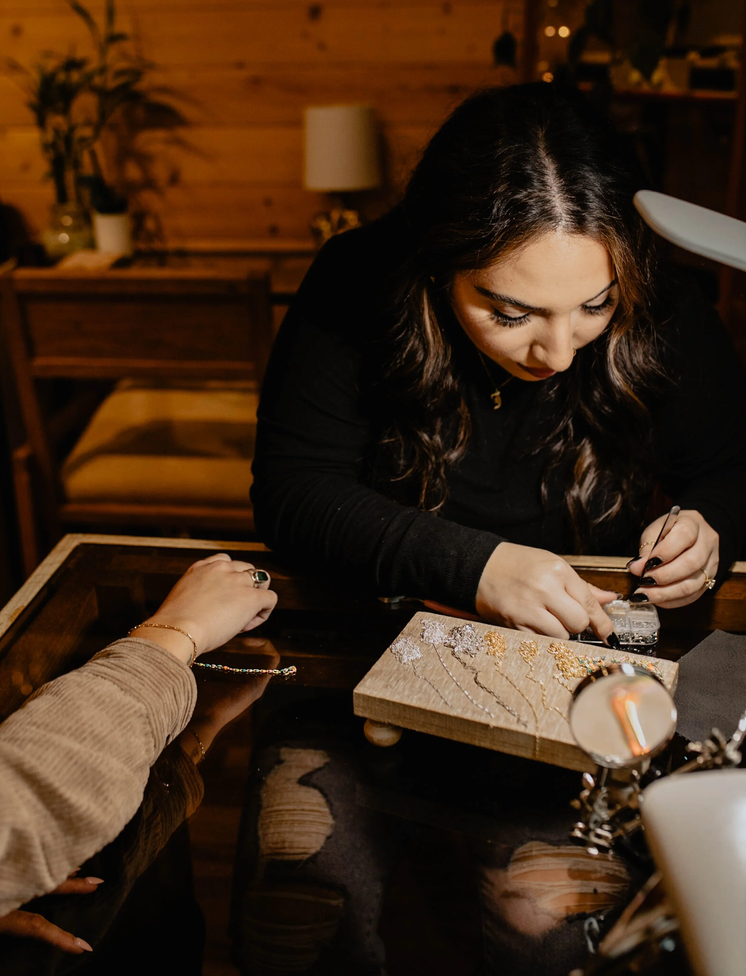 A woman with dark hair, wearing a black top, is working on a jewelry piece at a wooden table in a cozy, warmly lit room. Seattle professional head shot photography