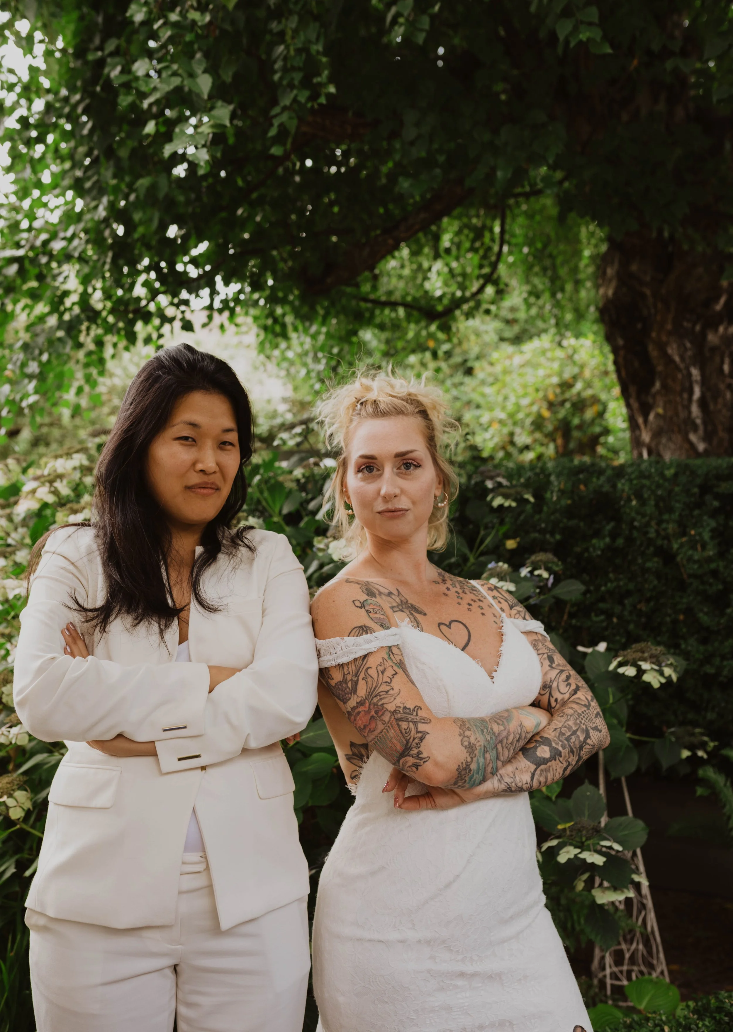 Two women standing outdoors with crossed arms, surrounded by green foliage and trees. One woman has dark hair, wearing a cream-colored suit, and the other has blond hair, tattoos, and is wearing a white dress.