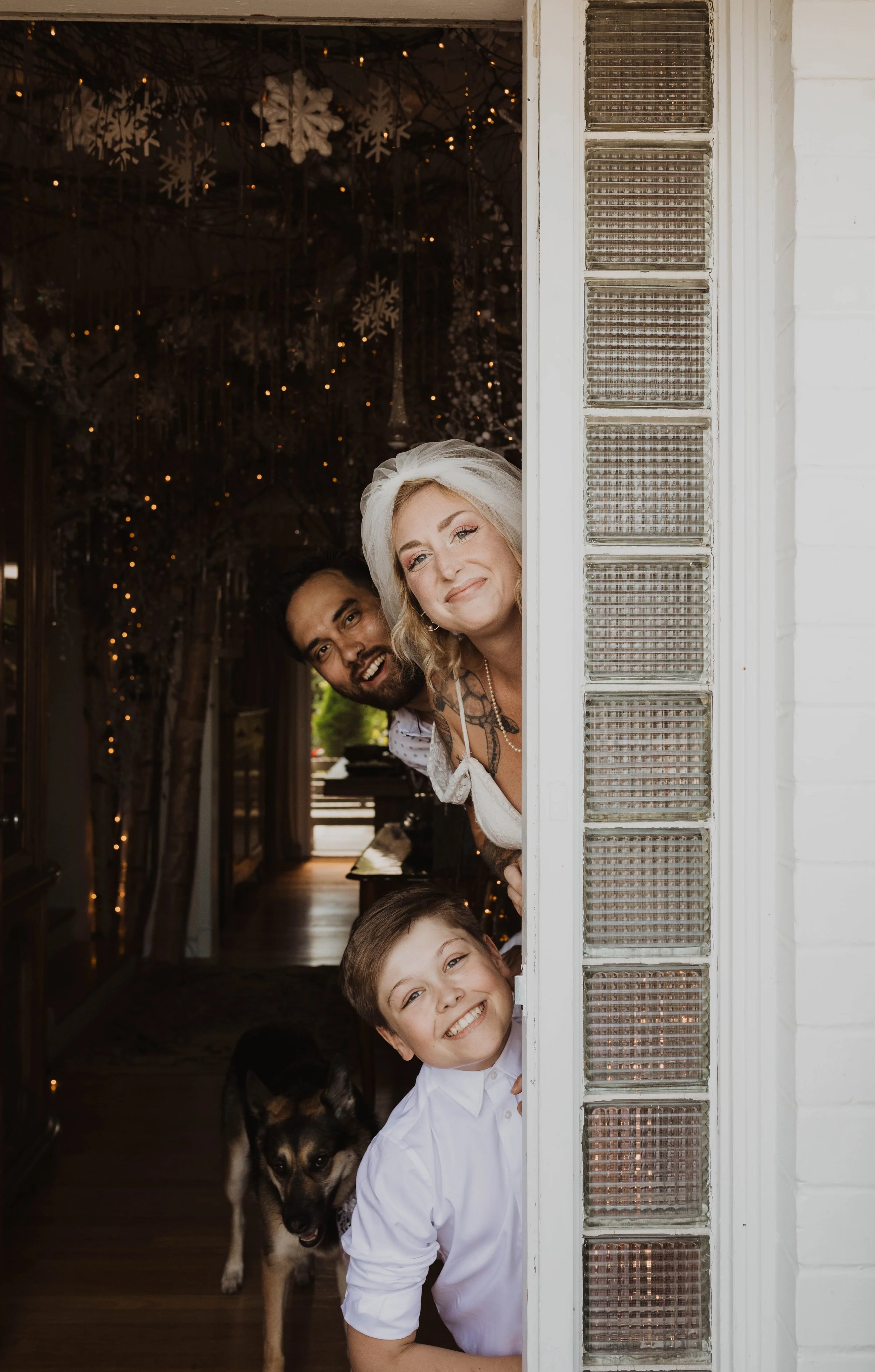A smiling woman with blonde hair, a man with dark hair and beard, and a boy with brown hair and a dog, all peeking through a door or window, with snowflake decorations and string lights inside Seattle, WA wedding photography.