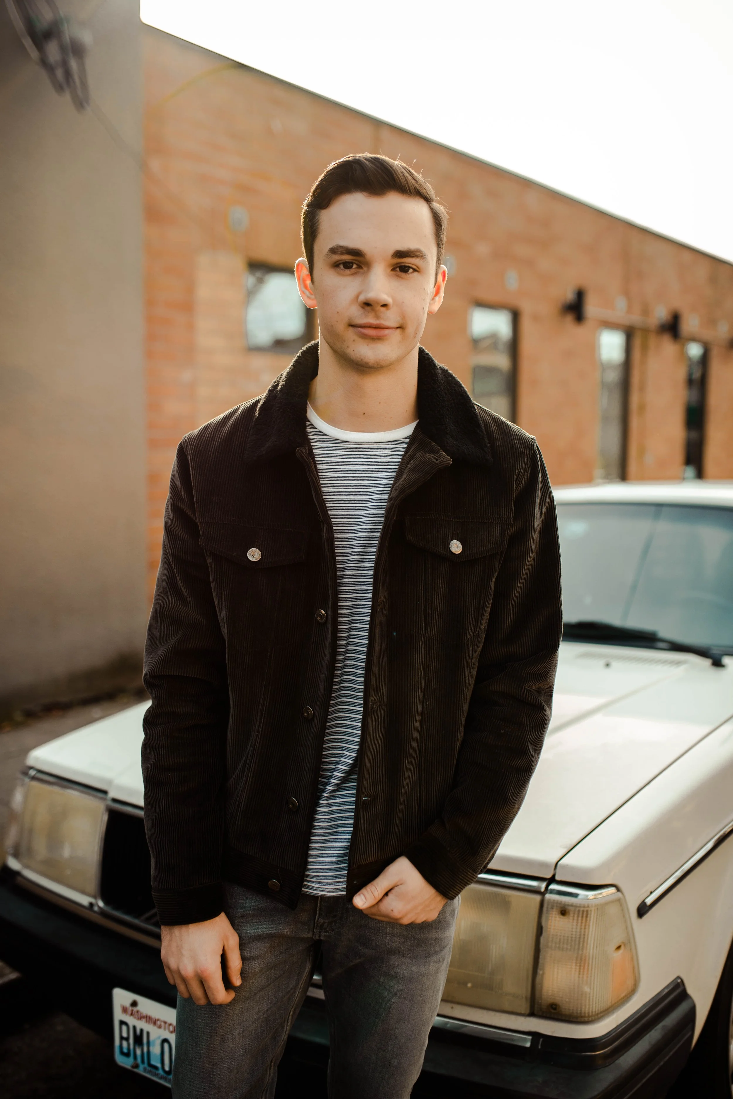 A young man with dark hair, wearing a black jacket and striped shirt, standing in front of a vintage white car parked beside a brick building. Seattle professional head shot photographer.