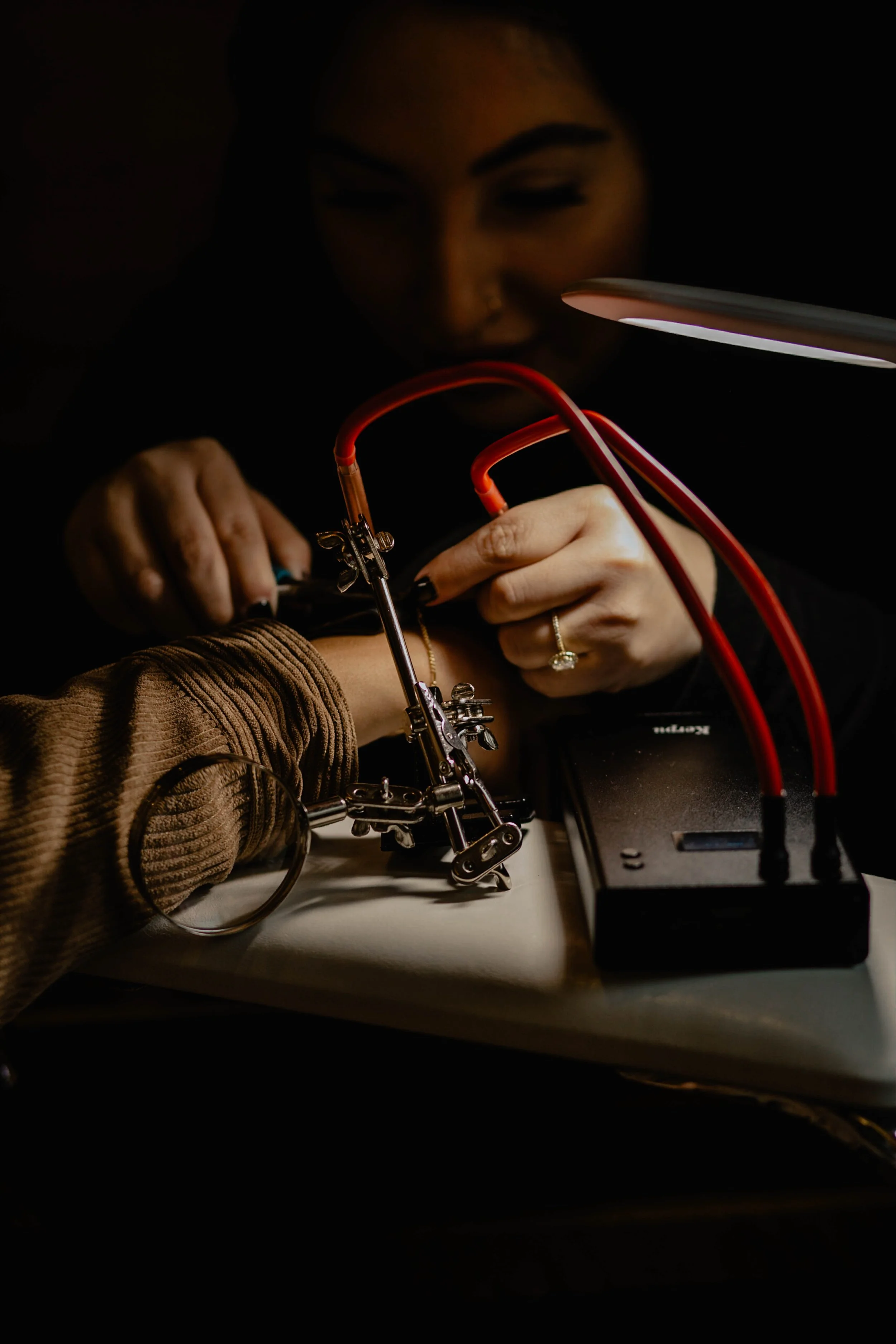 A woman is using a soldering iron on a person's arm, performing a tattoo. The scene is dimly lit, focusing on the tattooing process. Seattle professional head shot photography
