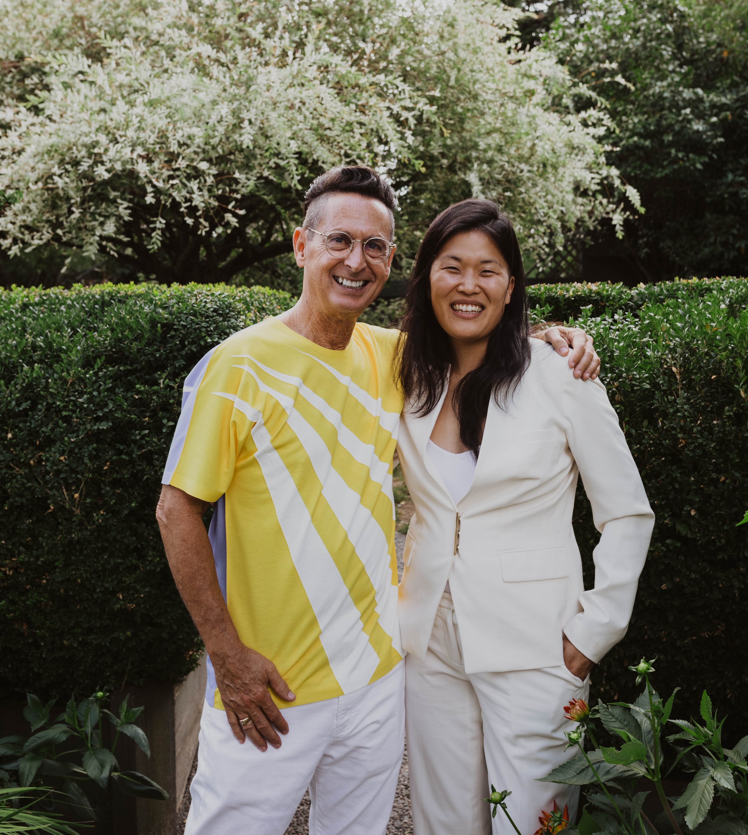 A man and woman standing outdoors, smiling, with the man having his arm around the woman's shoulders. They are surrounded by green bushes and trees. Seattle, WA wedding photography.