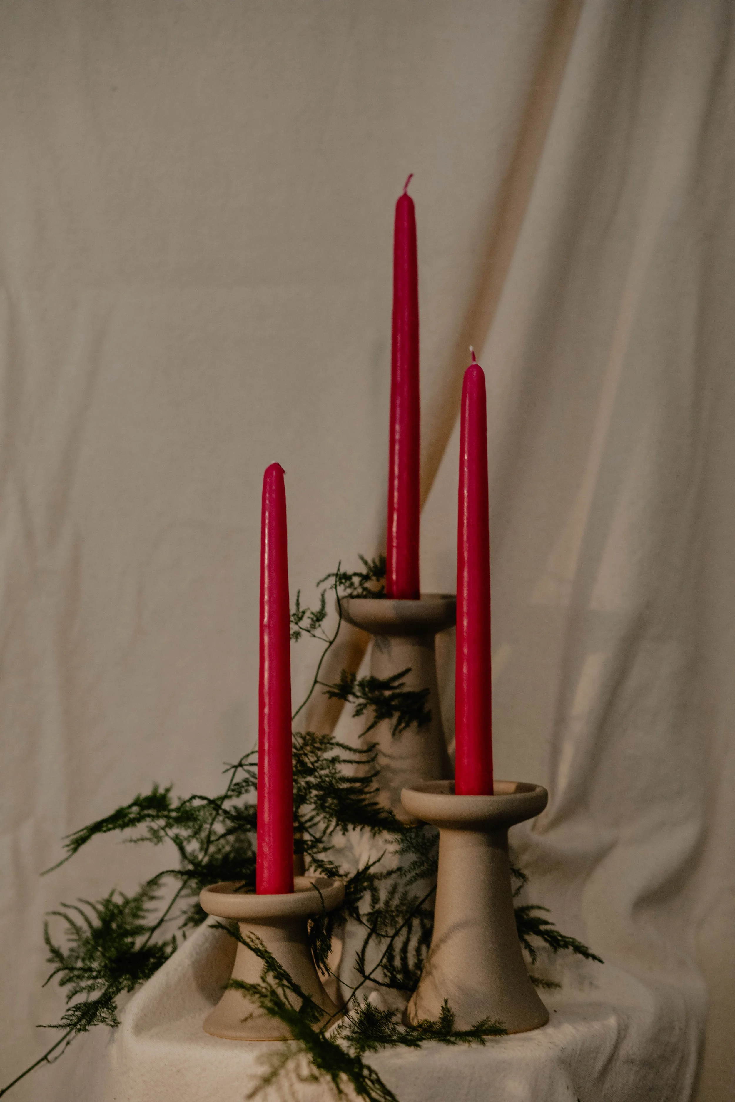 Three beige ceramic candlesticks with red tapered candles, surrounded by green foliage, on a white fabric surface.  Seattle product photography