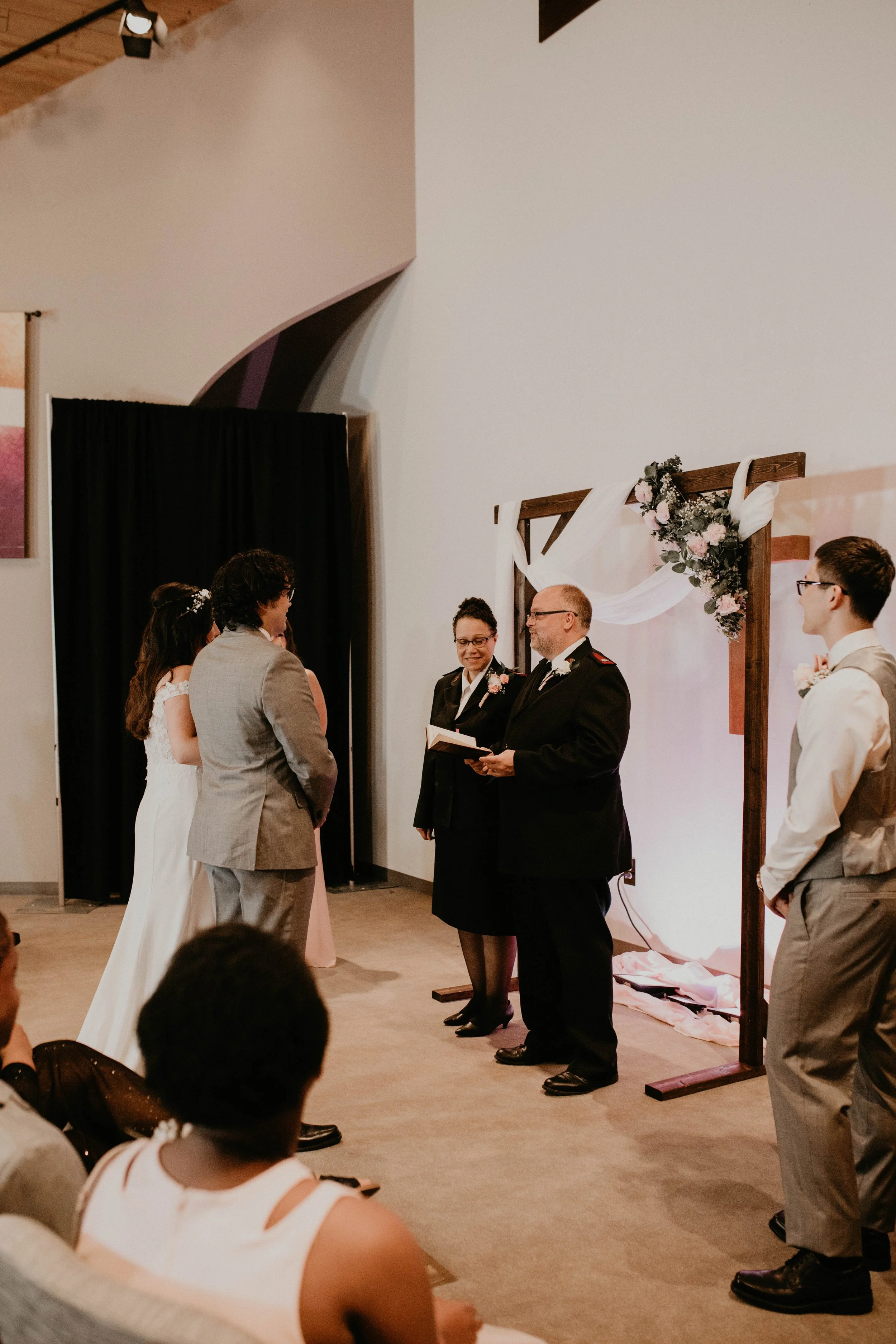 A wedding ceremony with the officiant and bride and groom standing facing each other, surrounded by wedding party members, with guests seated in front. The setting features a wooden arch decorated with flowers and draped fabric. Seattle, WA wedding p