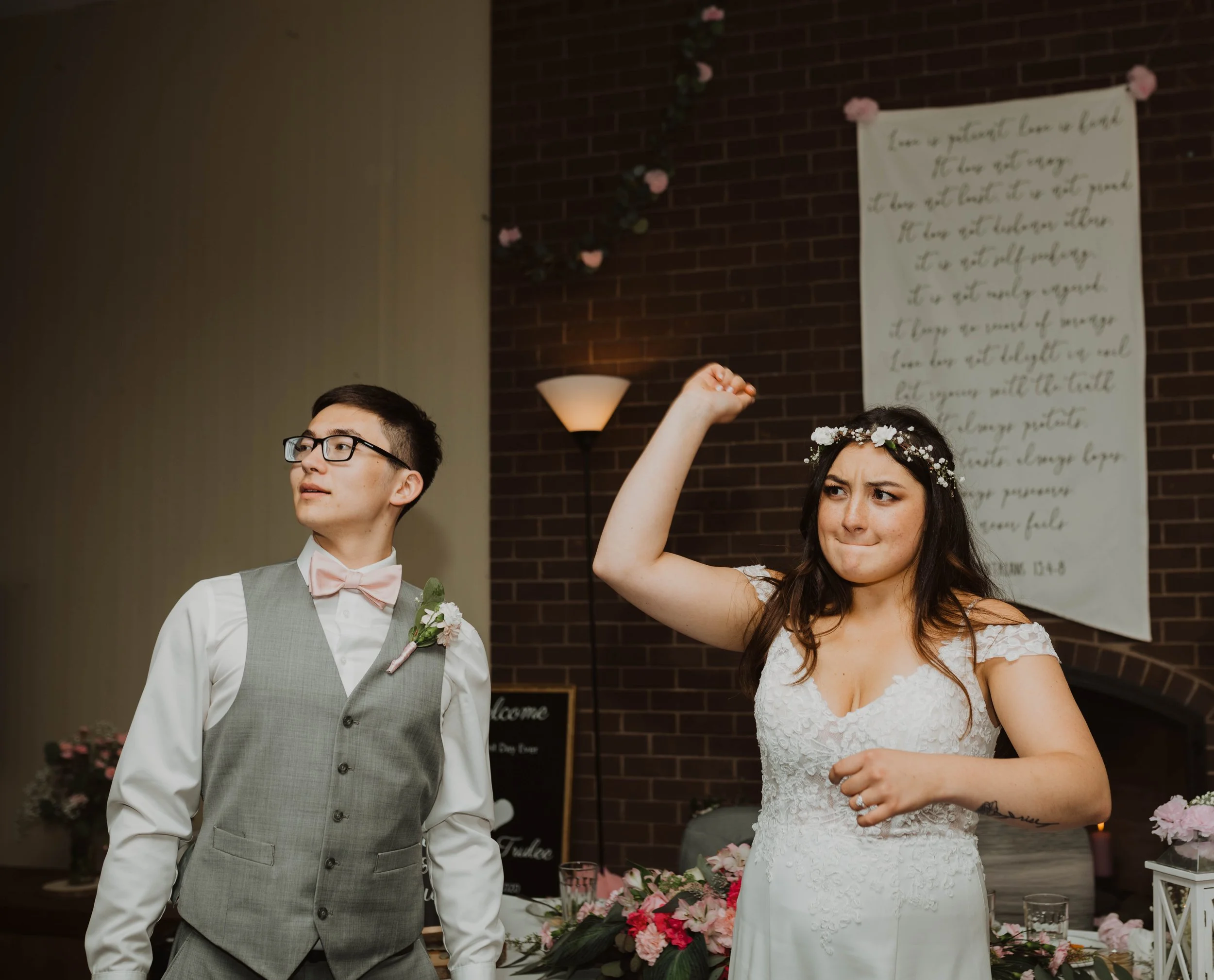 A bride and groom at their wedding reception, the bride is making a fist with her right hand and has a flower crown on her head, wearing a white lace wedding dress, while the groom is looking to the left, dressed in a gray vest and pink bow tie, with