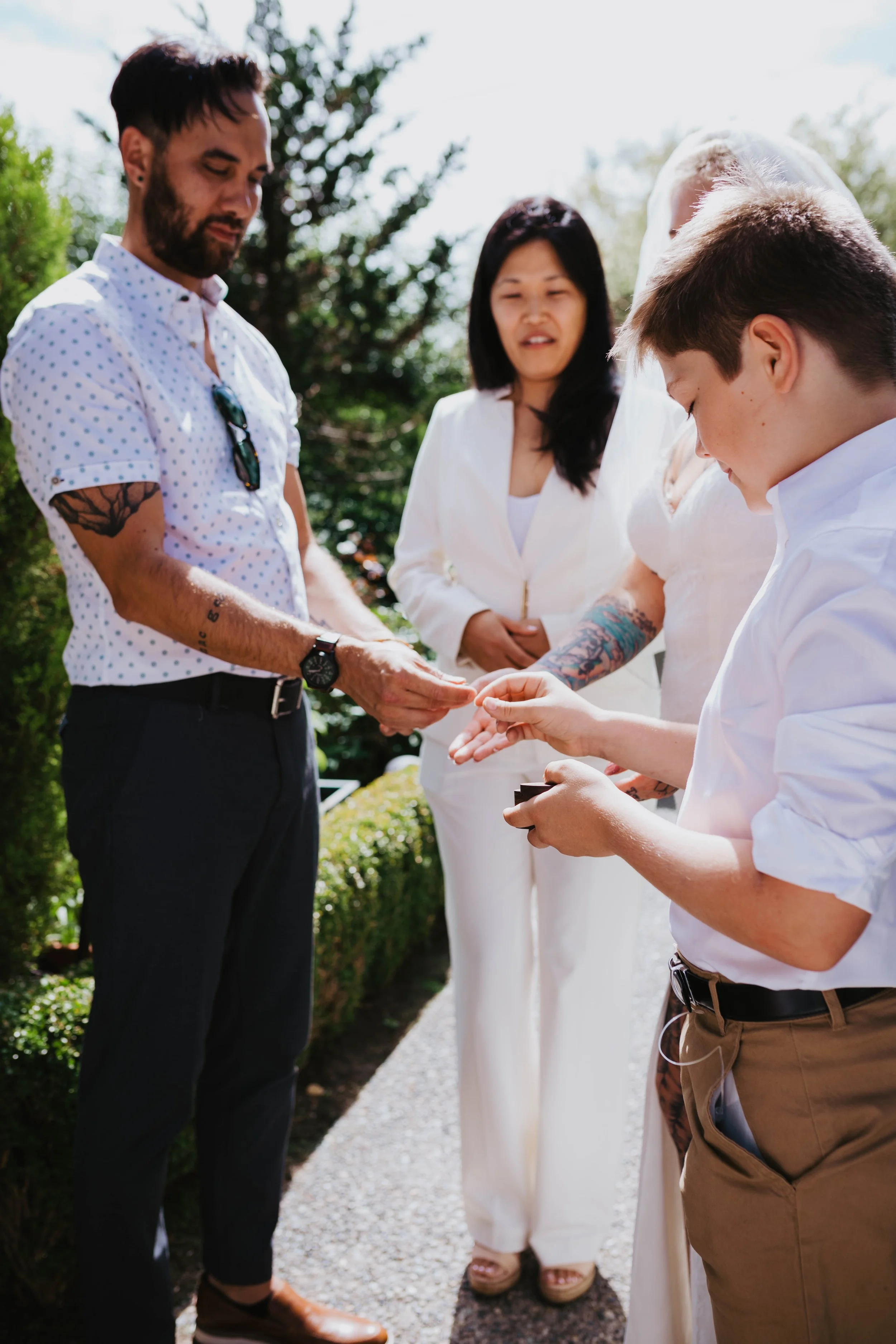 A wedding ceremony outdoors with four people, one of whom is a bride in a white dress and veil, and another is a groom, with two other individuals, exchanging rings. Seattle, WA wedding photography.