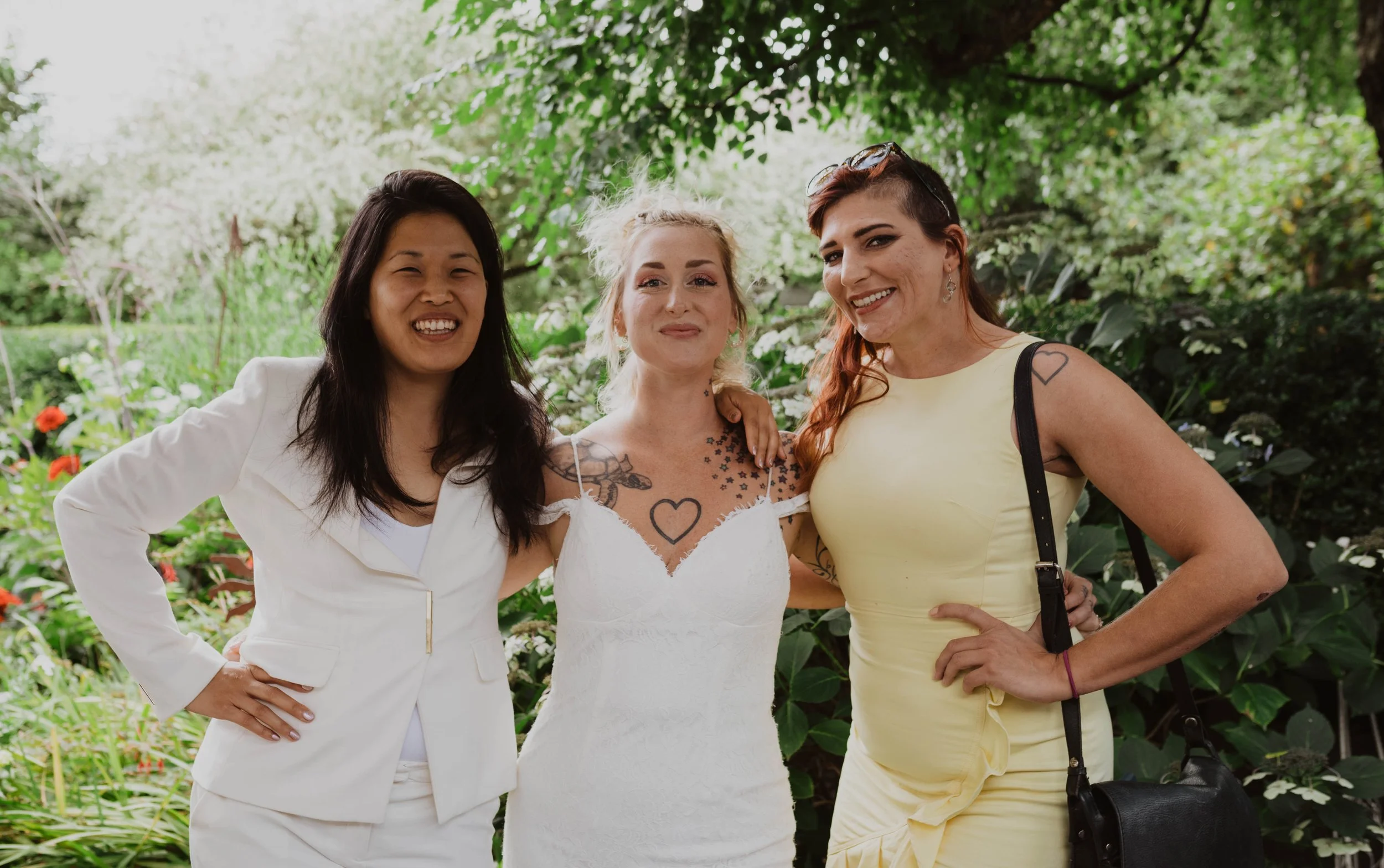 Three women smiling and posing together outdoors in a garden with green trees and flowers, one in a white suit, one in a white dress, and one in a yellow dress. Seattle, WA wedding photography.
