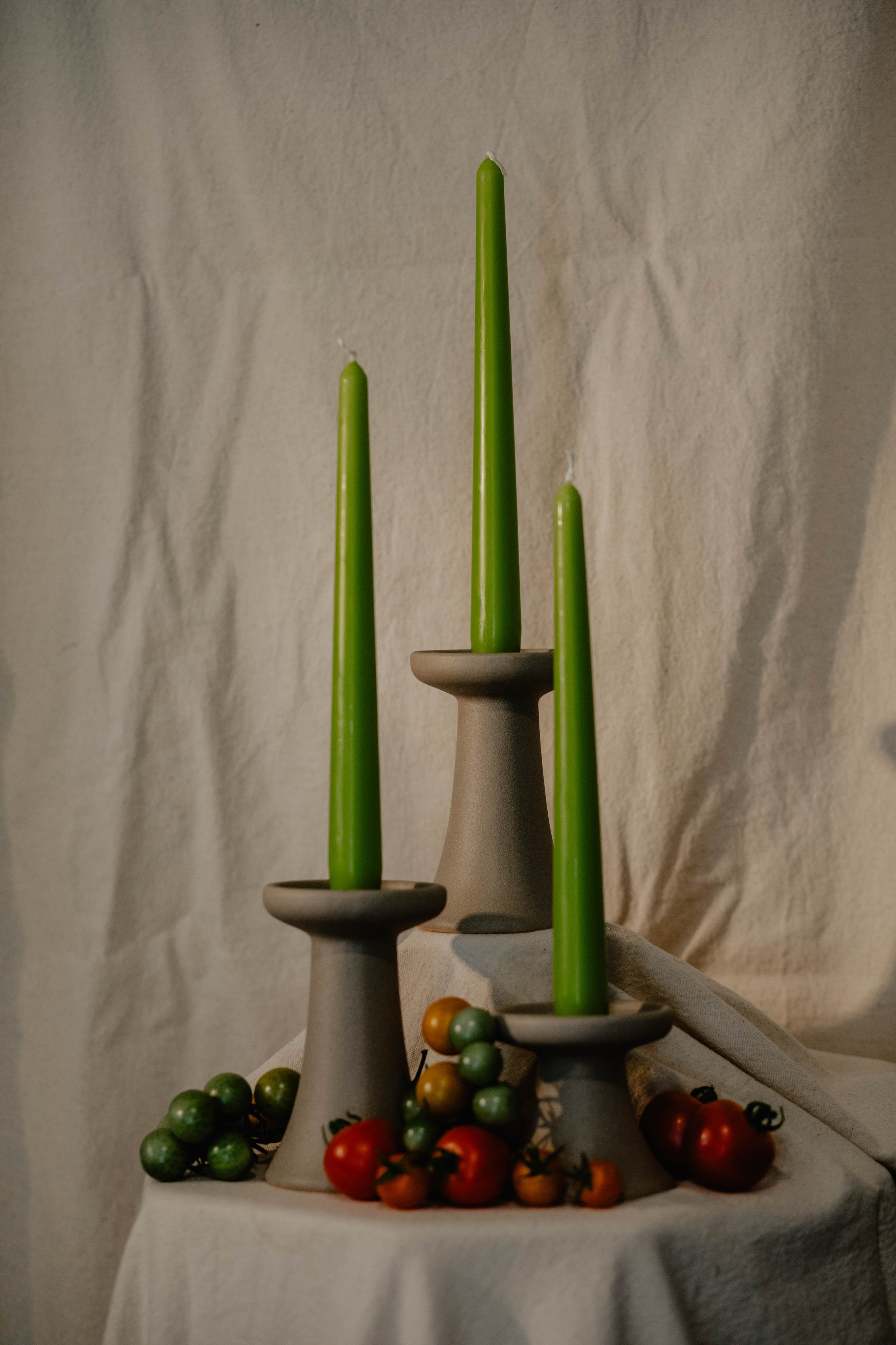 Three gray candle holders with tall green candles, surrounded by small tomatoes of various colors on a light-colored tablecloth. Seattle product photographer.