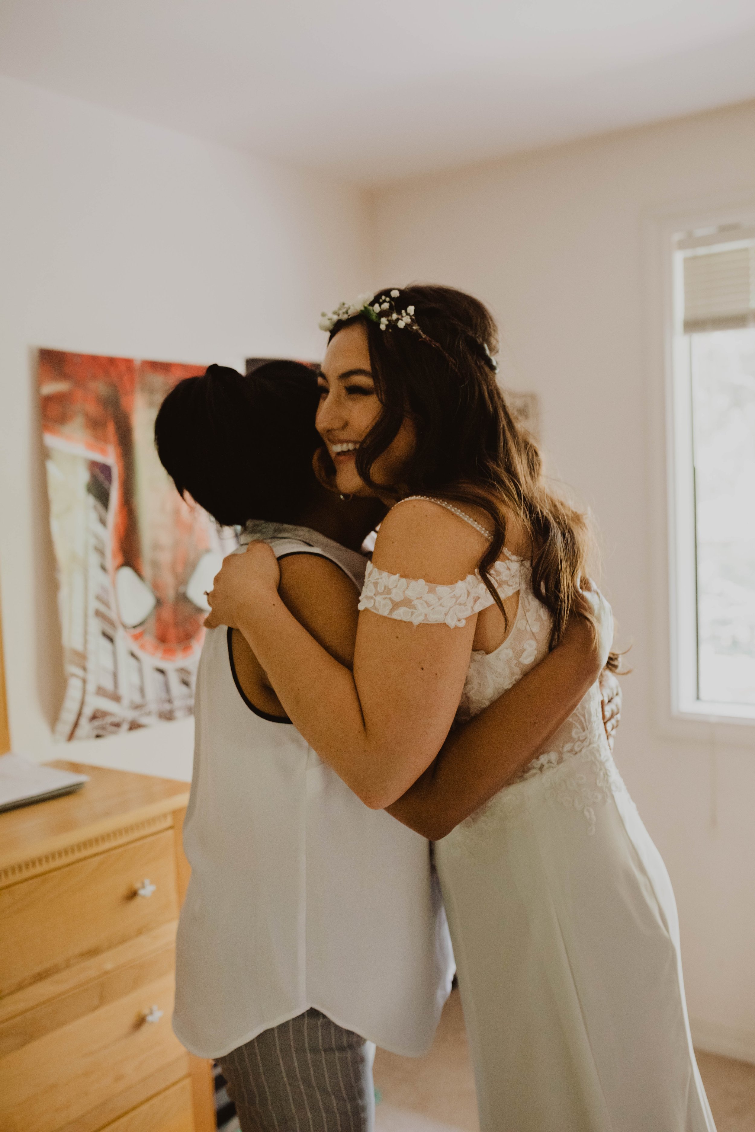 Two women embracing, one in a wedding dress, smiling, in a bright room with a window and a wooden dresser. Seattle, WA wedding photography.