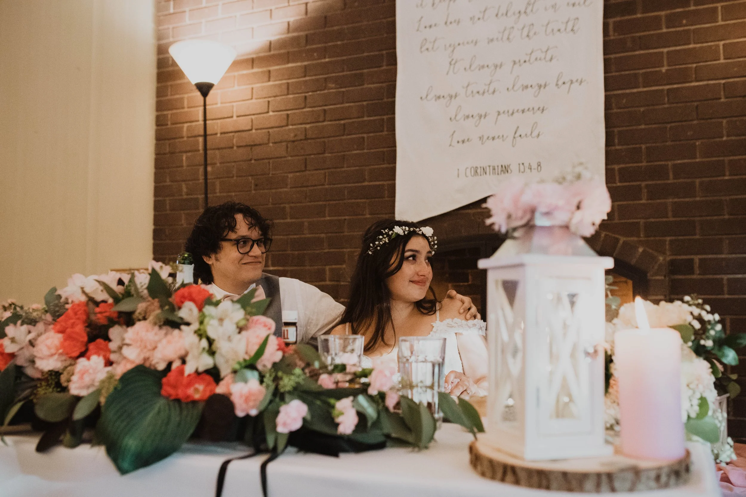 A bride and groom sitting at a wedding reception table decorated with pink and white flowers and candles, with a brick wall and a white banner with cursive writing in the background. Seattle, WA wedding photography.