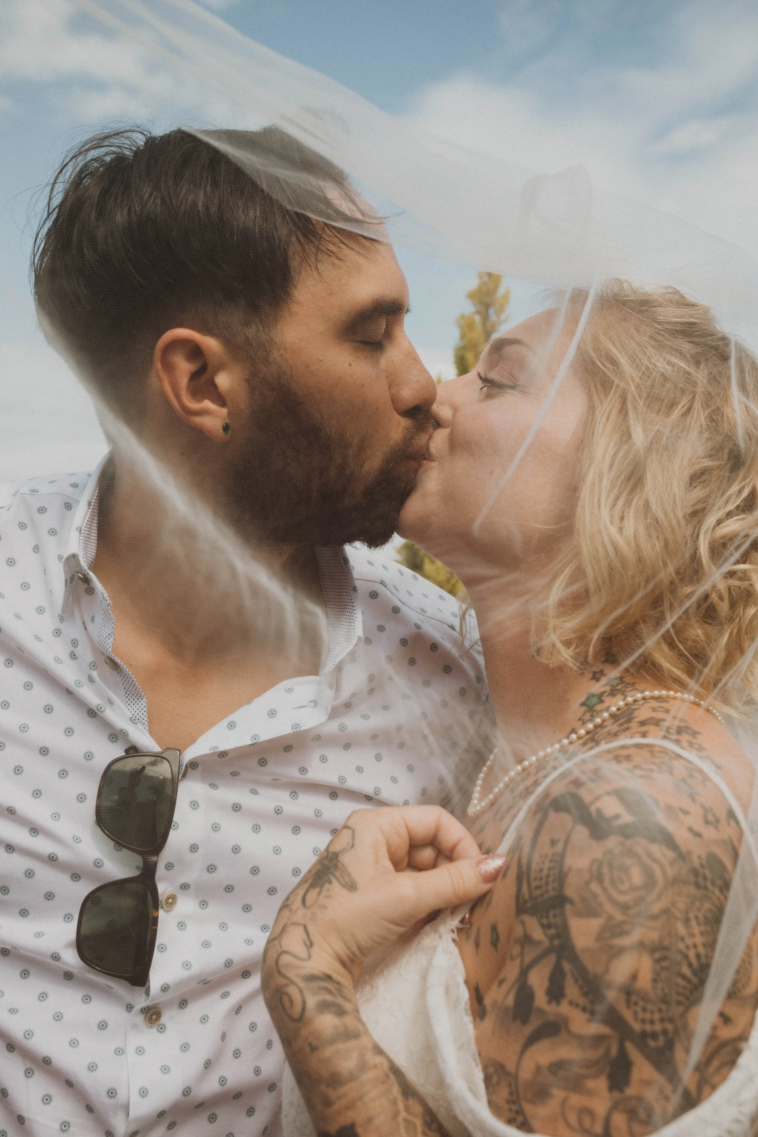 A couple sharing a kiss under a clear sky, with the woman having tattoos and wearing a pearl necklace, and the man with sunglasses hanging from his shirt. Seattle, WA wedding photography.