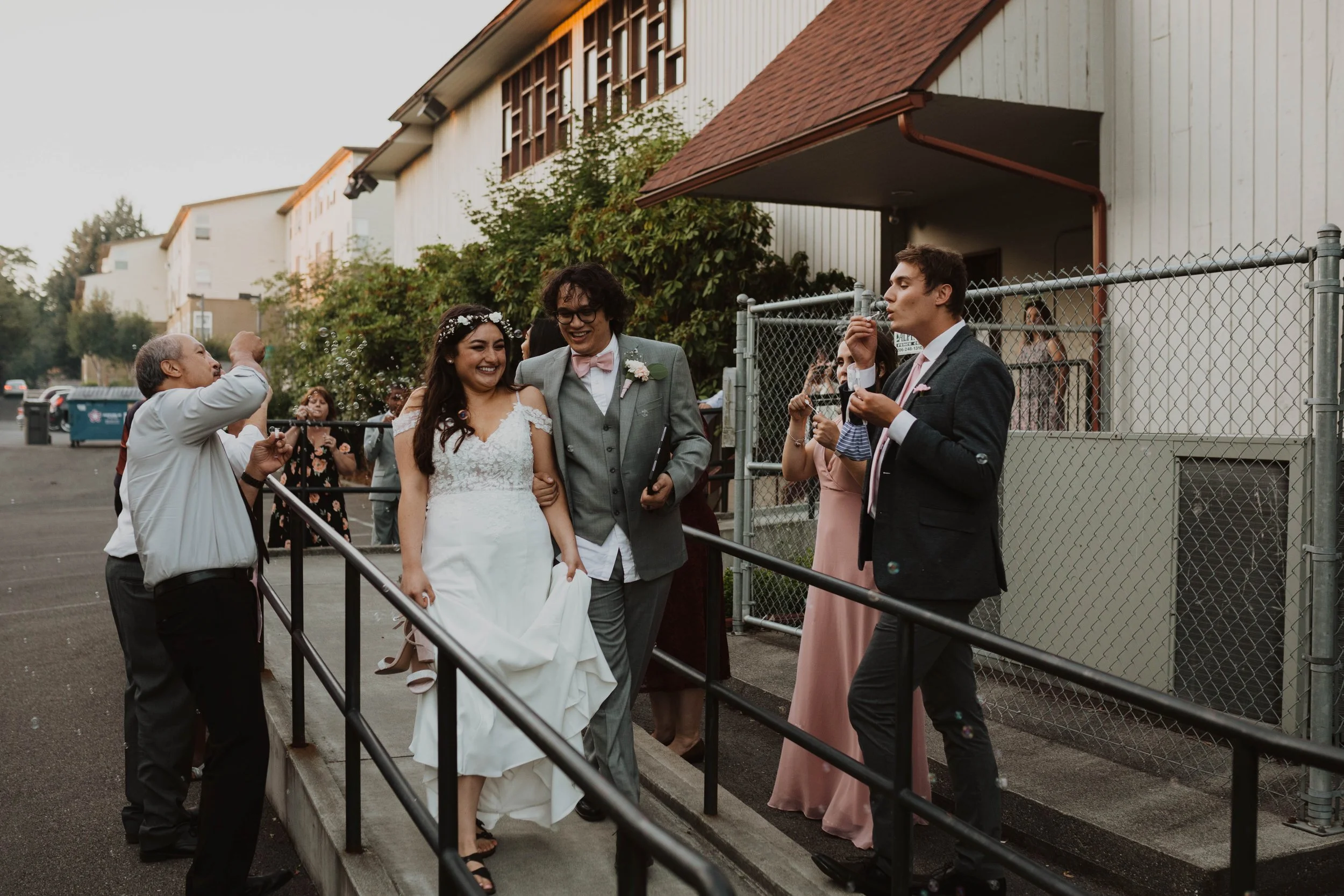 Wedding couple walking outside, bride in white wedding dress and groom in gray suit with pink bow tie, surrounded by friends and guests taking photos and blowing bubbles. Seattle, WA wedding photography.