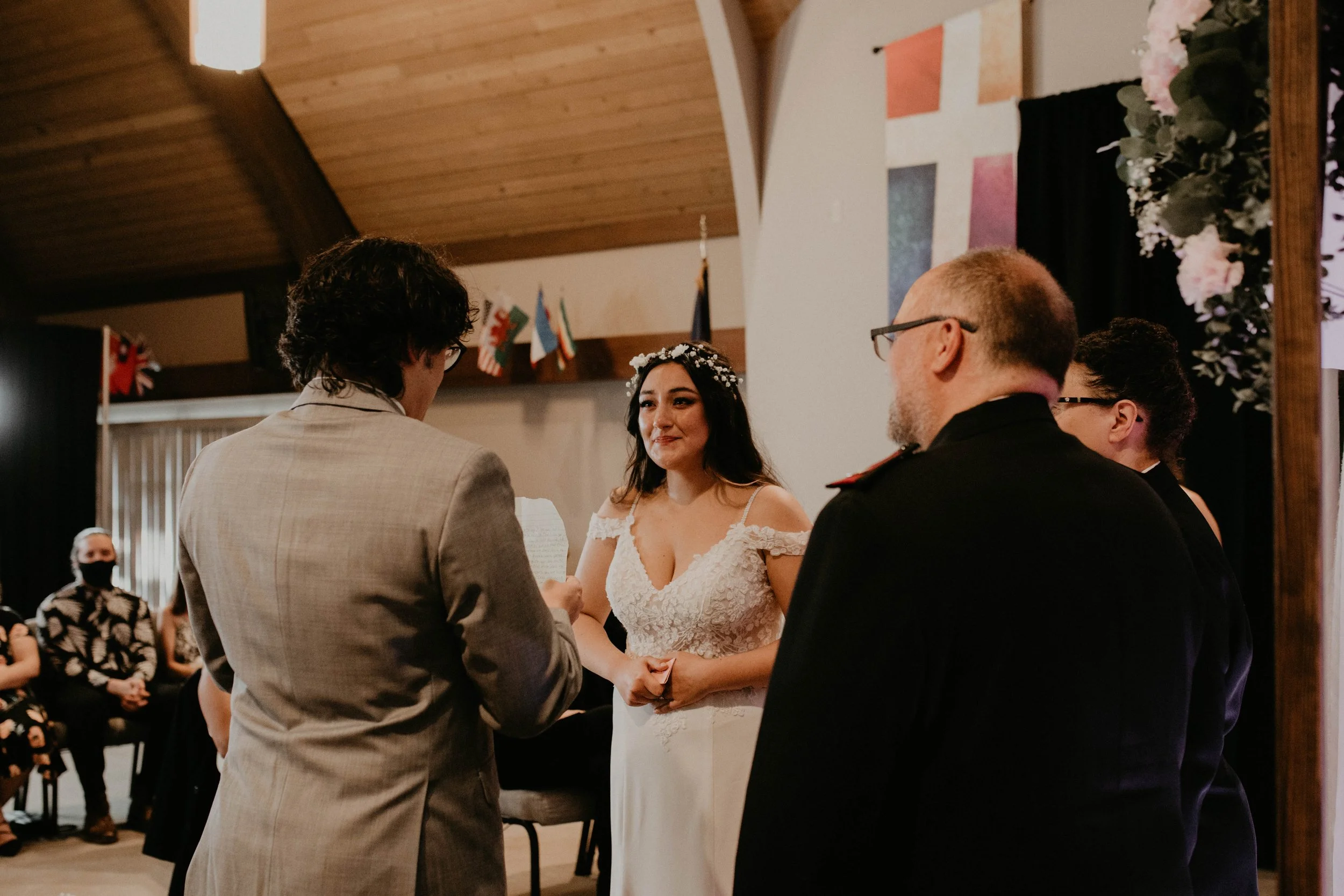 A bride with dark hair and a floral crown stands with a groom during a wedding ceremony indoors. The bride is smiling and holding hands with the groom, who is facing away from the camera. Two officiants are standing with the bride and groom, and gues
