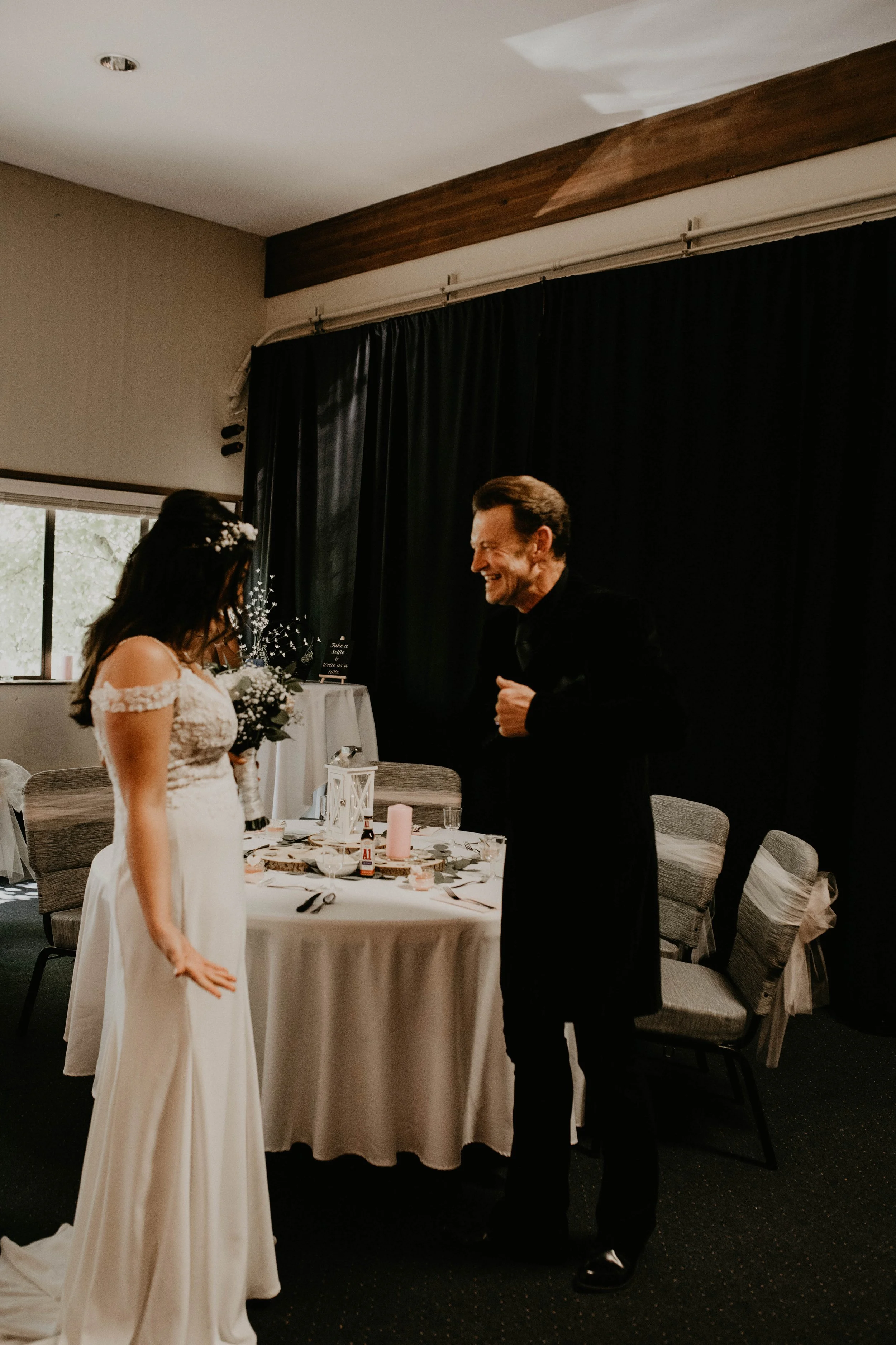 A bride with dark hair in a white wedding dress holding a bouquet, standing in front of a smiling man in a black suit during a wedding reception in a decorated indoor venue. Seattle, WA wedding photography.