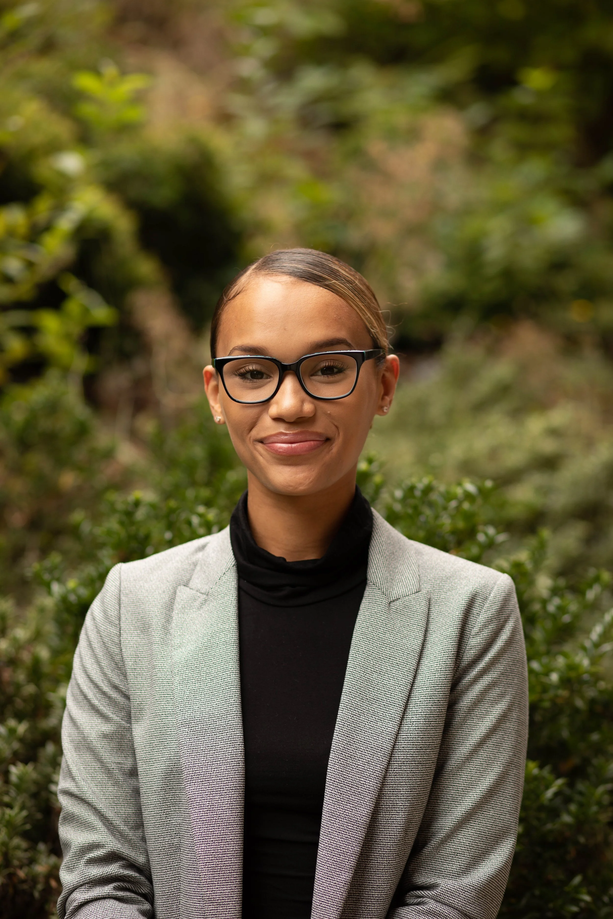A young woman with glasses, smiling, standing outdoors with greenery in the background. Seattle professional head shot photography